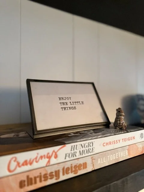 A framed sign on a shelf reads "ENJOY THE LITTLE THINGS." Next to it is a ceramic figure of a sitting dog or cat, and below are books titled "Cravings," "Hungry For More," and "All Together."