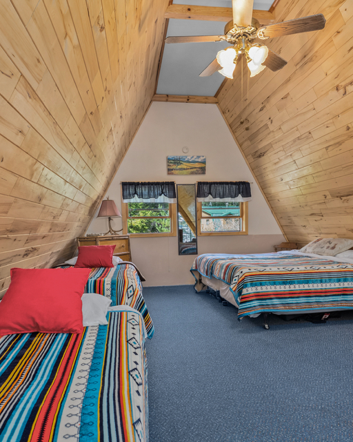 Room with wood-paneled sloped ceiling, two beds with colorful striped bedspreads, and a ceiling fan.