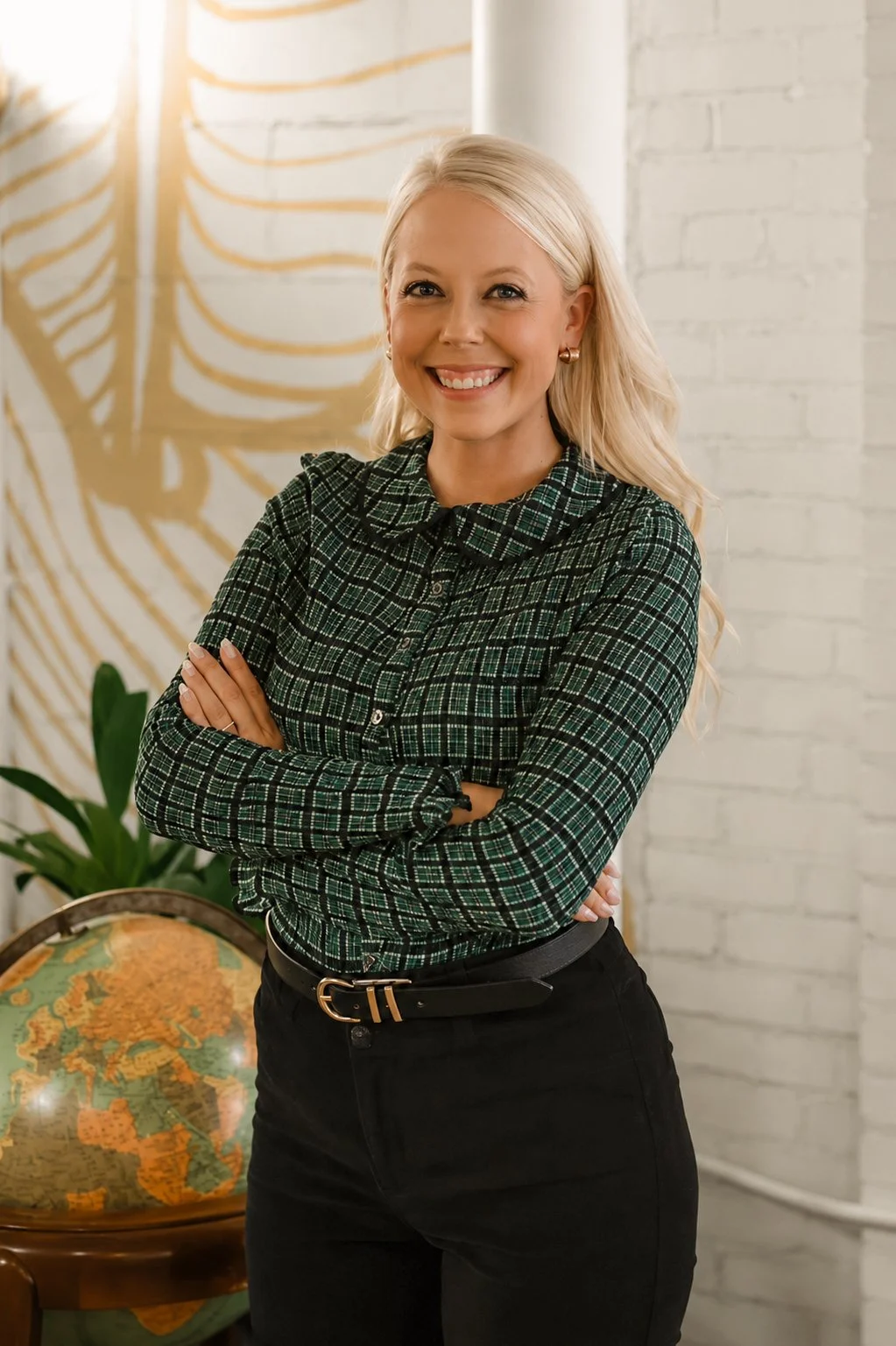 A woman with blonde hair, smiling, wearing a green checkered shirt and black pants, standing with arms crossed in a bright room with a world globe, green plant, and abstract wall art in the background.