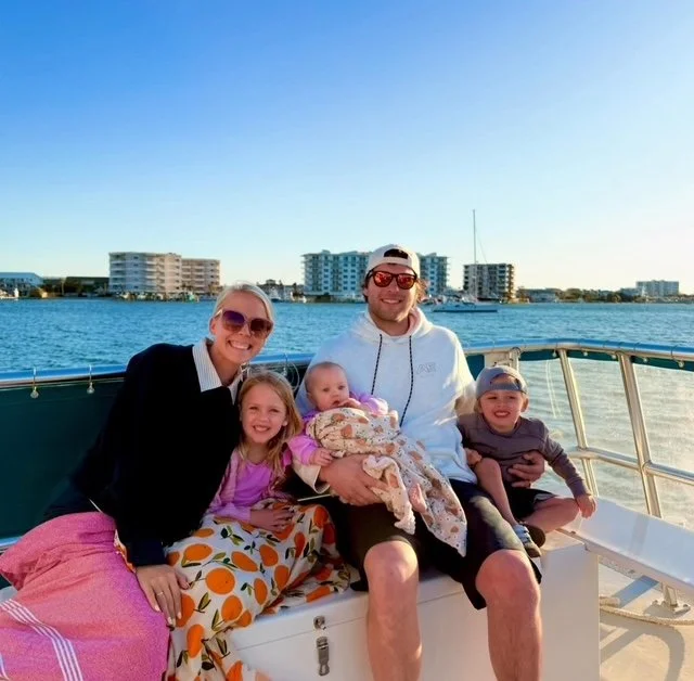 A family of five sitting on a boat docked on a waterway with buildings in the background. The family includes a woman wearing sunglasses, a man with sunglasses and a cap, and three children, two girls and a boy, smiling at the camera.