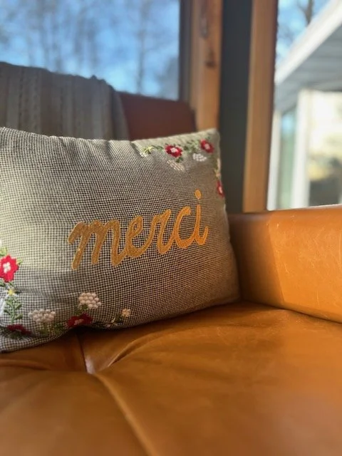 Decorative pillow with embroidered pink and white flowers and the word 'merci' in wooden cursive on a leather couch.