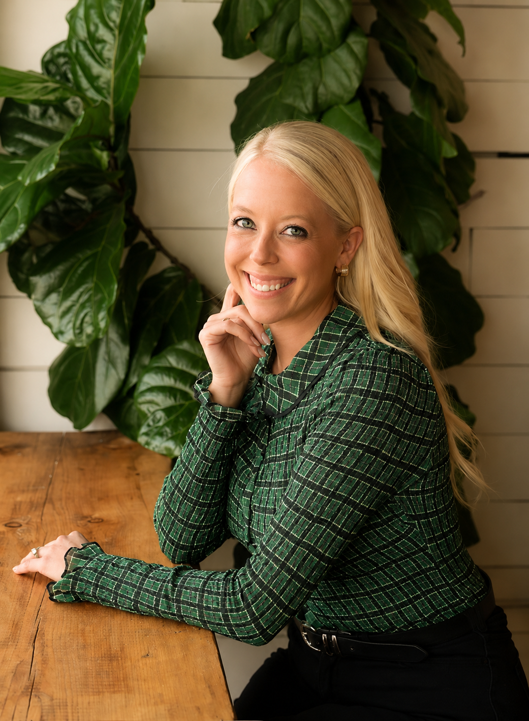 A smiling blonde woman in a green plaid shirt sitting at a wooden table with a large green leafy plant behind her.