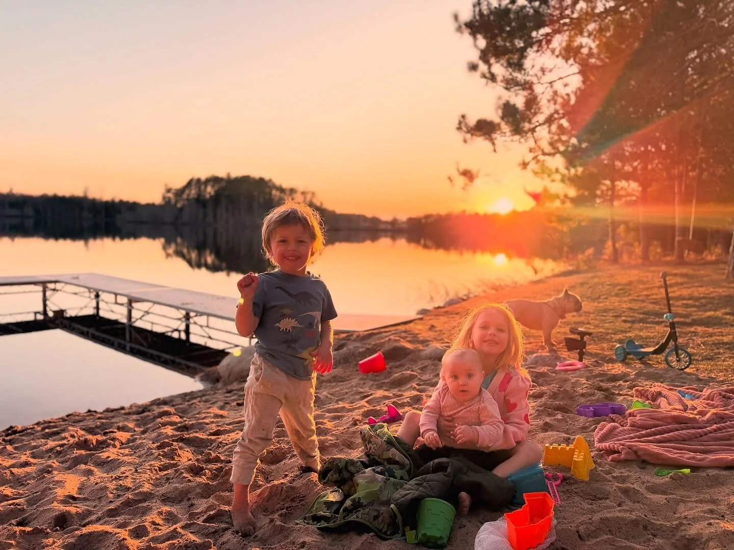 Three children playing on a beach at sunset, with toys scattered around, a dog walking nearby, and a calm lake in the background.
