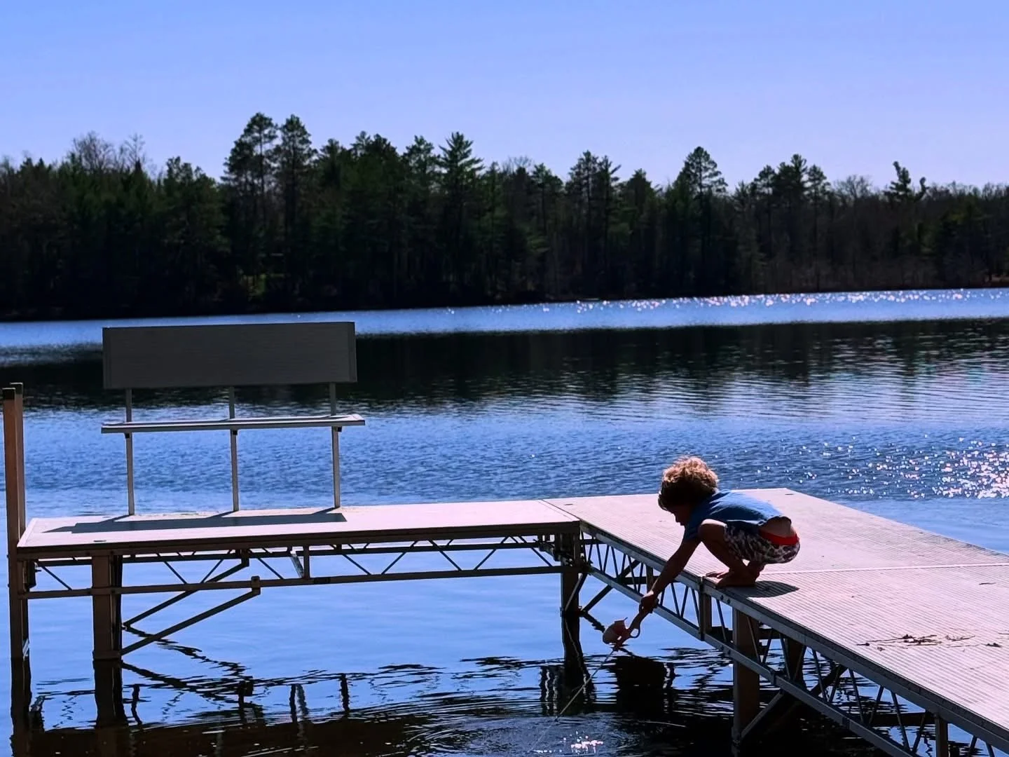 A child playing at the edge of a dock on a lake, with a forested shoreline in the background. The child is bending down, reaching into the water.