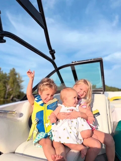 Three children sitting on a boat seat under a partially open canopy, with a blue sky and green trees in the background, enjoying a sunny day.