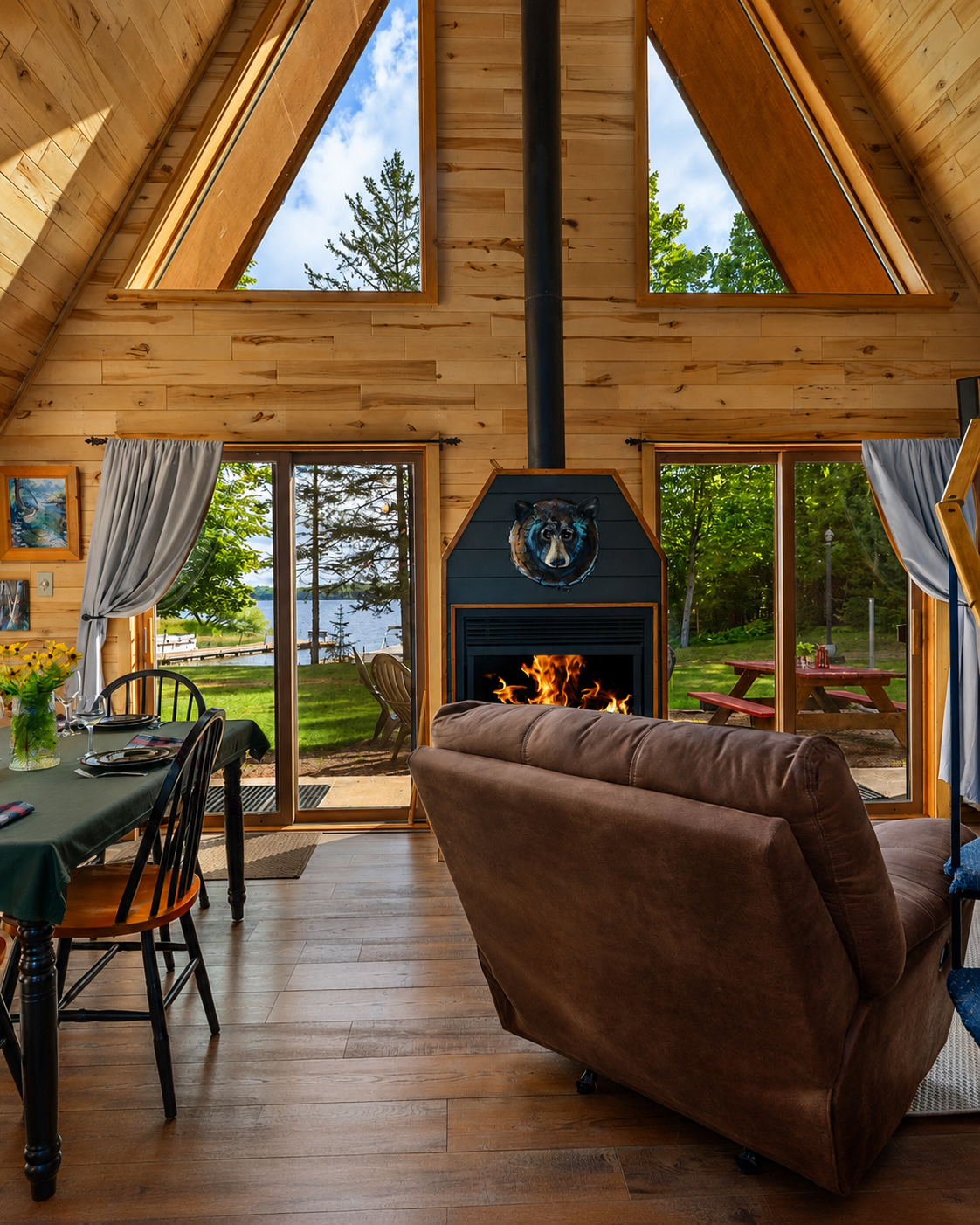 The interior of a wooden cabin living room with a high vaulted ceiling, large triangular windows showing trees and a lake outside, a fireplace with a bear's head sculpture above it, a brown couch in front of the fireplace, a dining table with chairs and tableware, and sliding glass doors with curtains leading to a deck with picnic tables.