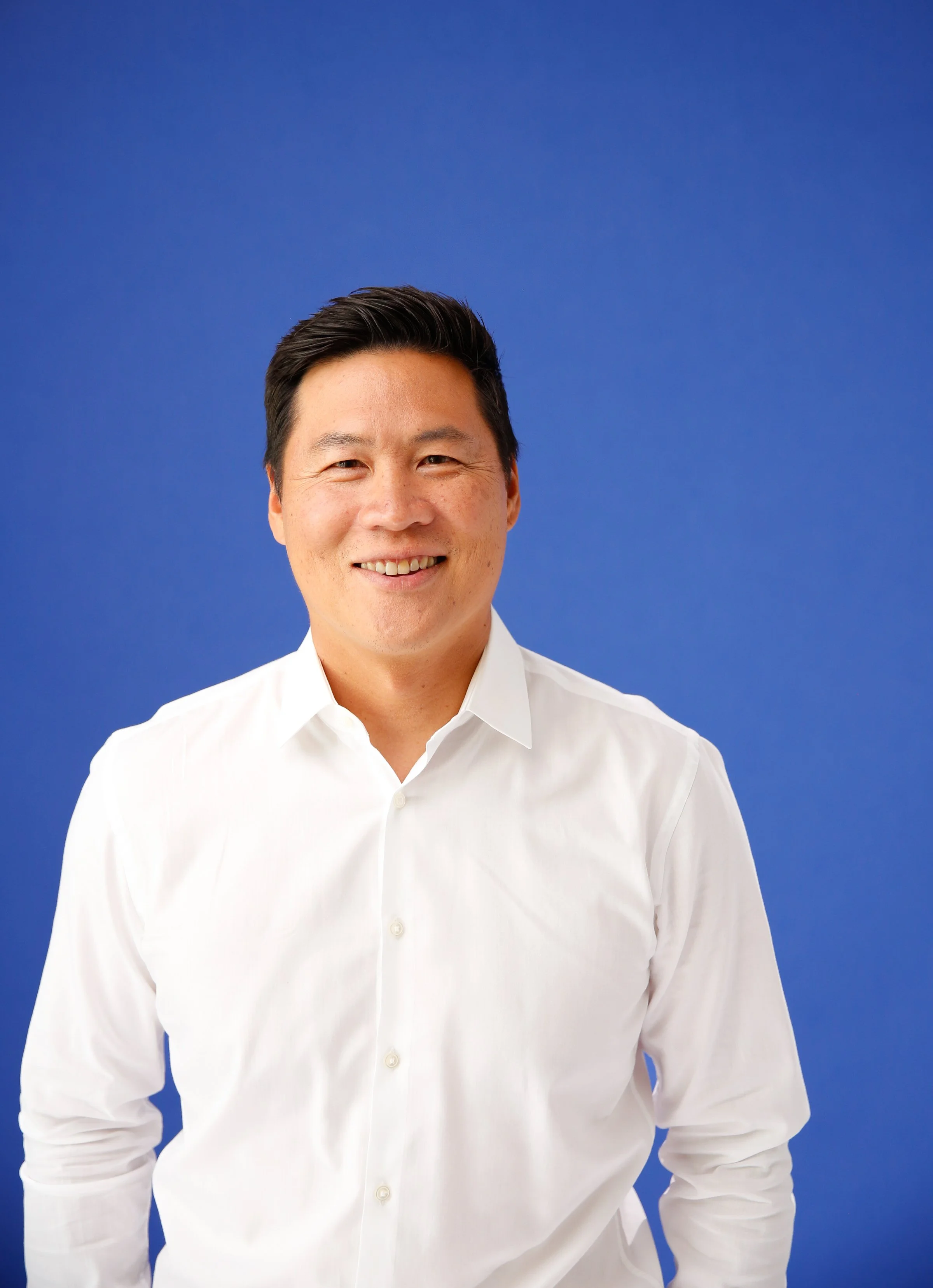 Headshot of a smiling man with dark hair, wearing a white shirt, against a solid blue background.