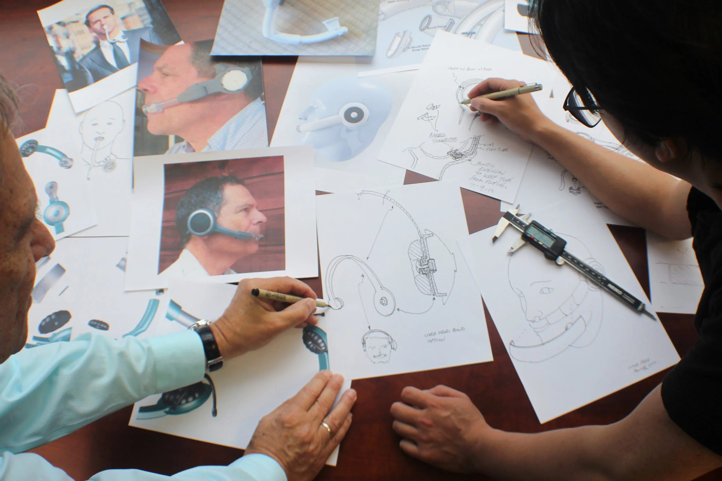 Group of people working on sketches and diagrams of medical devices, including headsets and breathing masks, with printed photos and technical drawings scattered on the table.