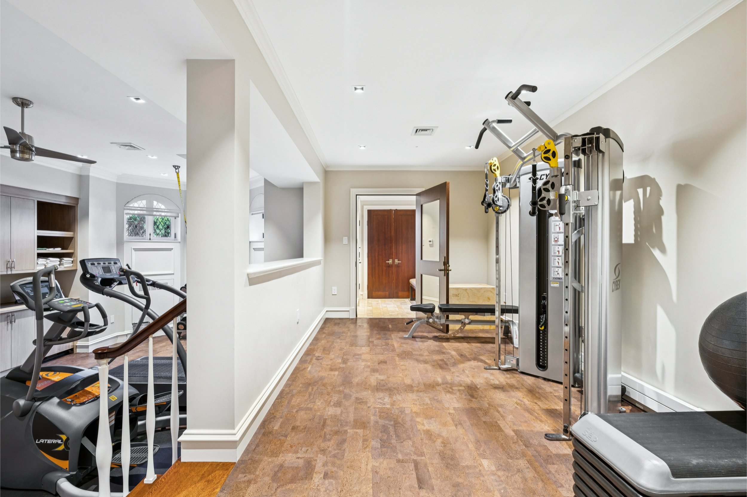 Home gym with various exercise equipment including treadmills, elliptical machines, weight lifting machine, and balance ball in a well-lit room with hardwood floors and white walls.