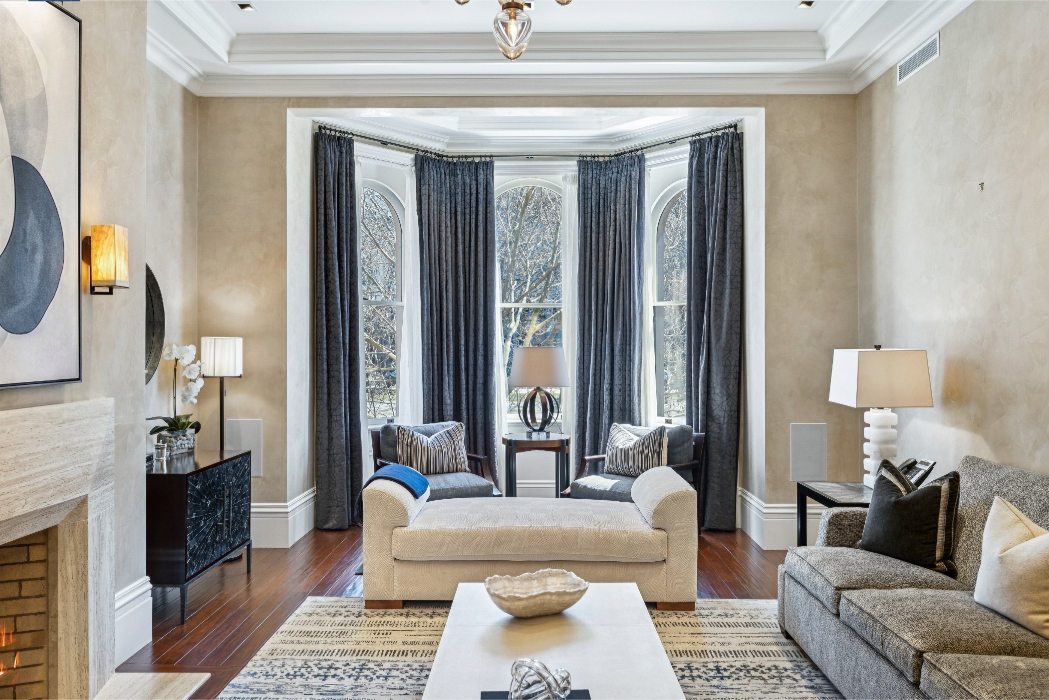 Living room with large bay windows with trees outside, beige and gray furniture, a coffee table, and various lamps.