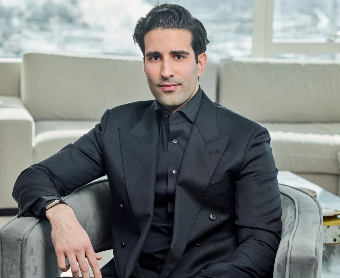 A man in a black suit sitting on a gray armchair in a modern indoor space with large windows and light-colored furniture.