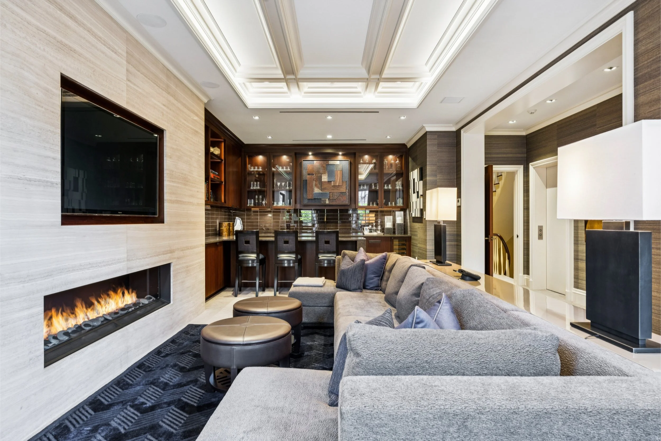 Living room with modern design, featuring a large gray sectional sofa, a fireplace, a flat-screen TV, a bar with seating, and decorative artwork on the walls.
