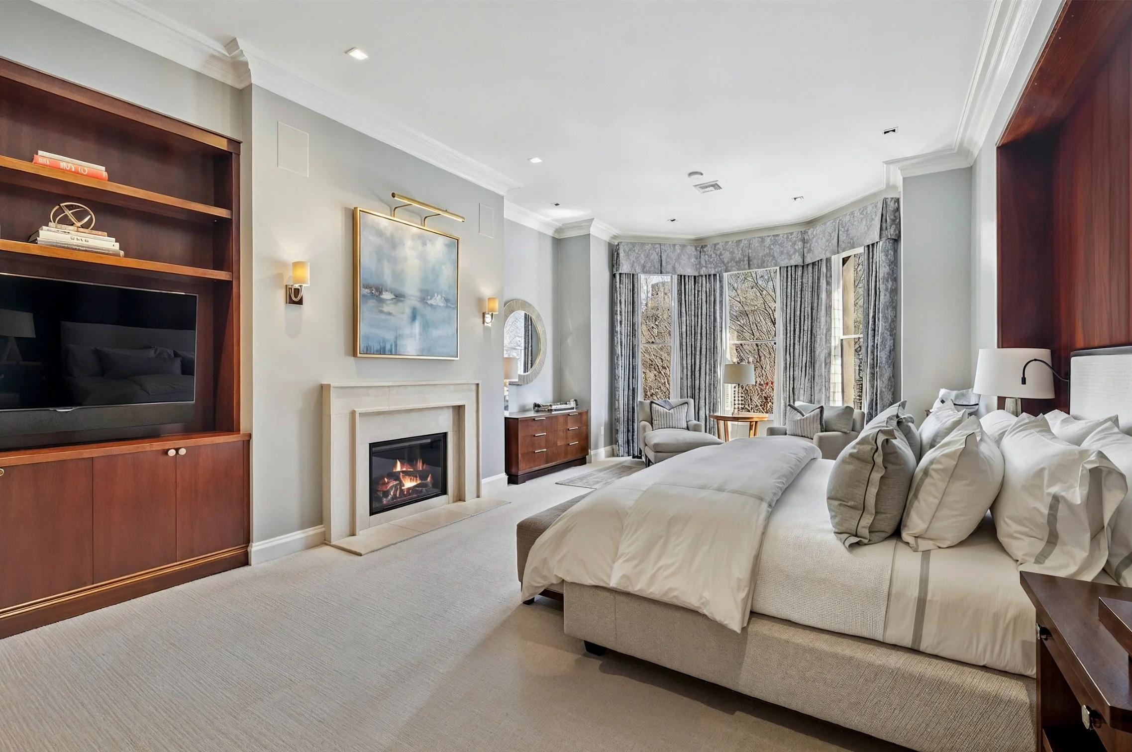 A spacious hotel room with a large bed, sitting area, fireplace, and large window with curtains showing an outdoor view of trees.