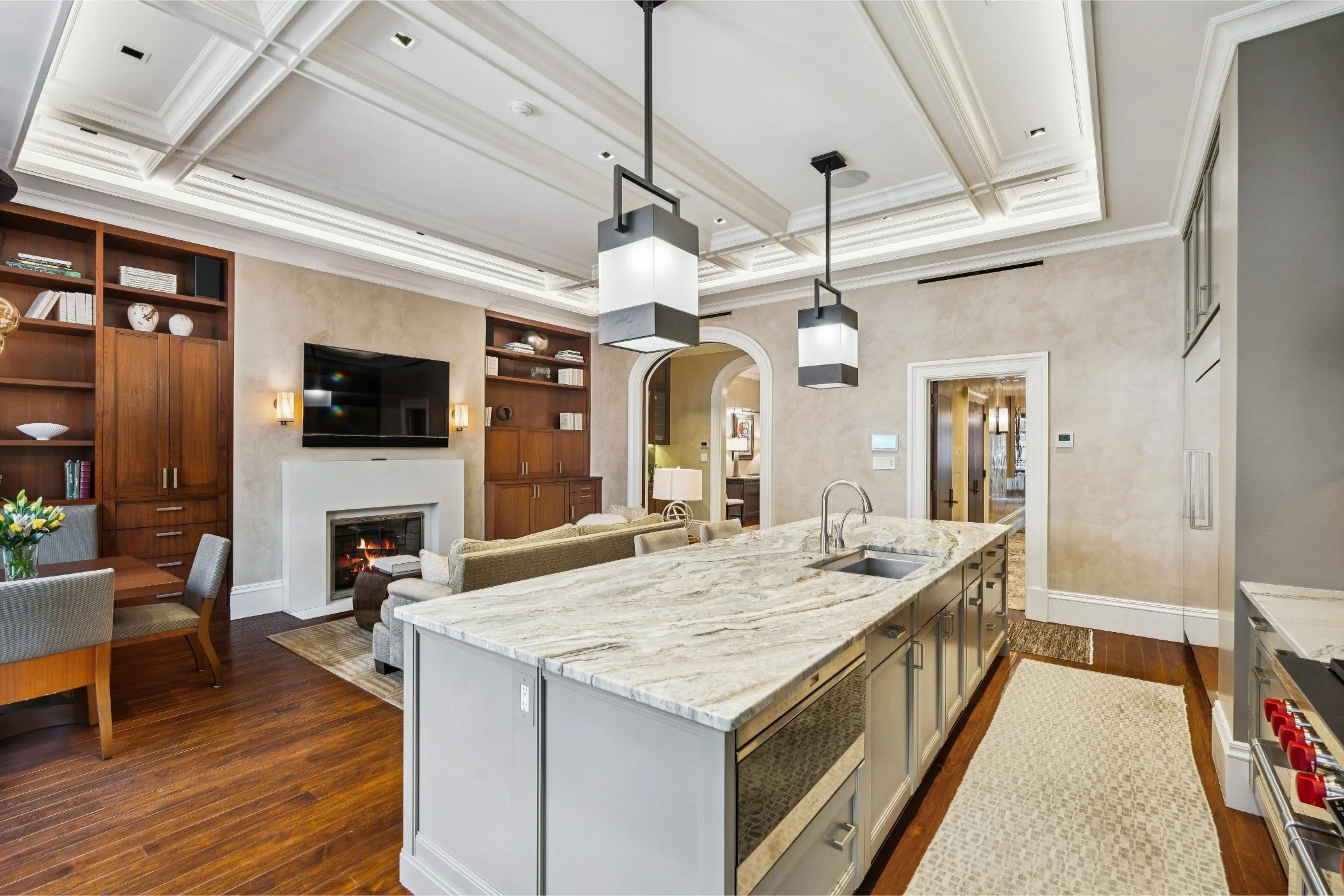 Modern open-concept living room and kitchen with white coffered ceiling, pendant lights, built-in wooden shelving, a fireplace, and a kitchen island with marble countertop and sink.