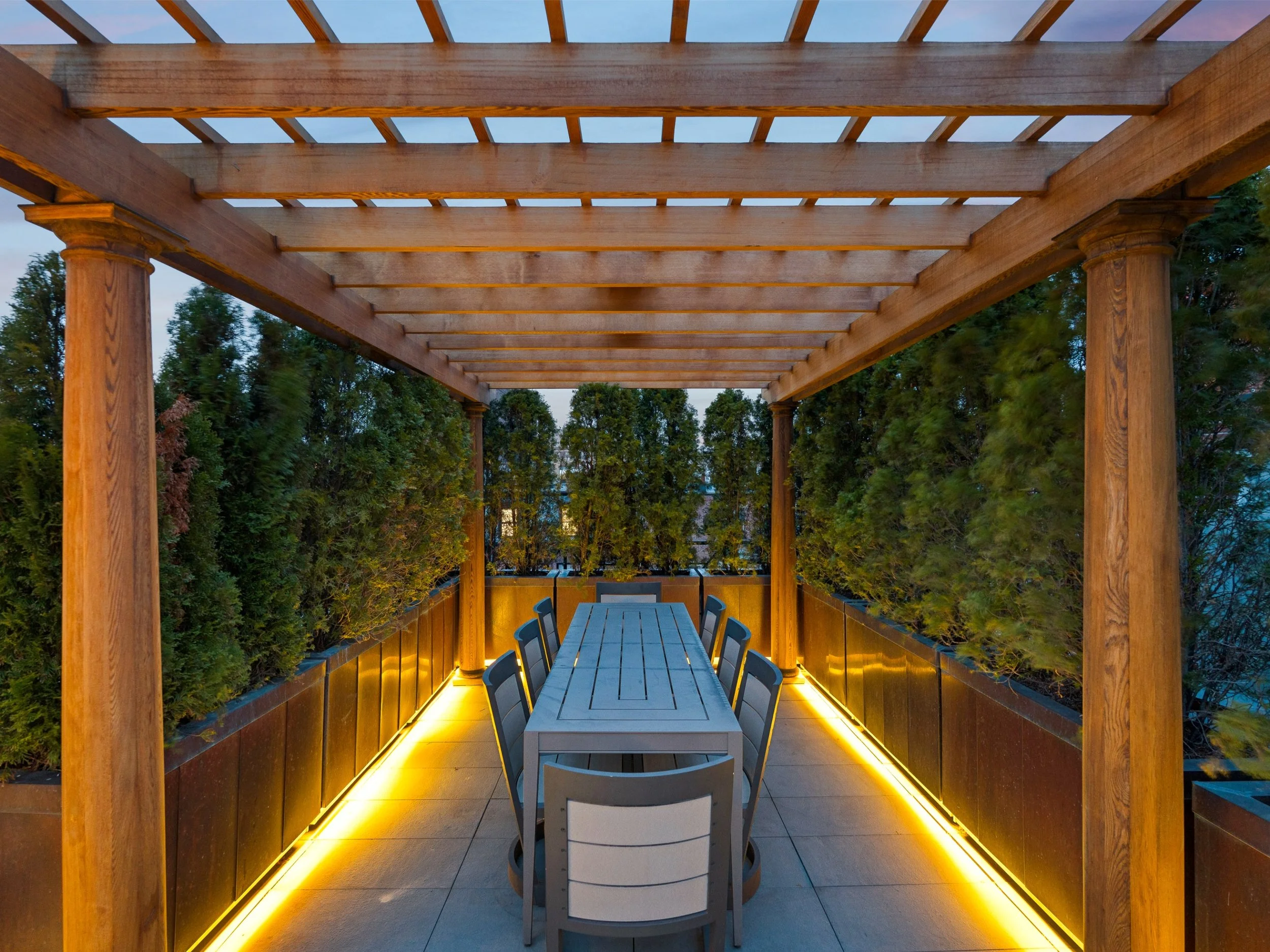 Outdoor dining area under a wooden pergola with a long table, multiple chairs, surrounded by greenery, lit with warm yellow lighting at dusk.