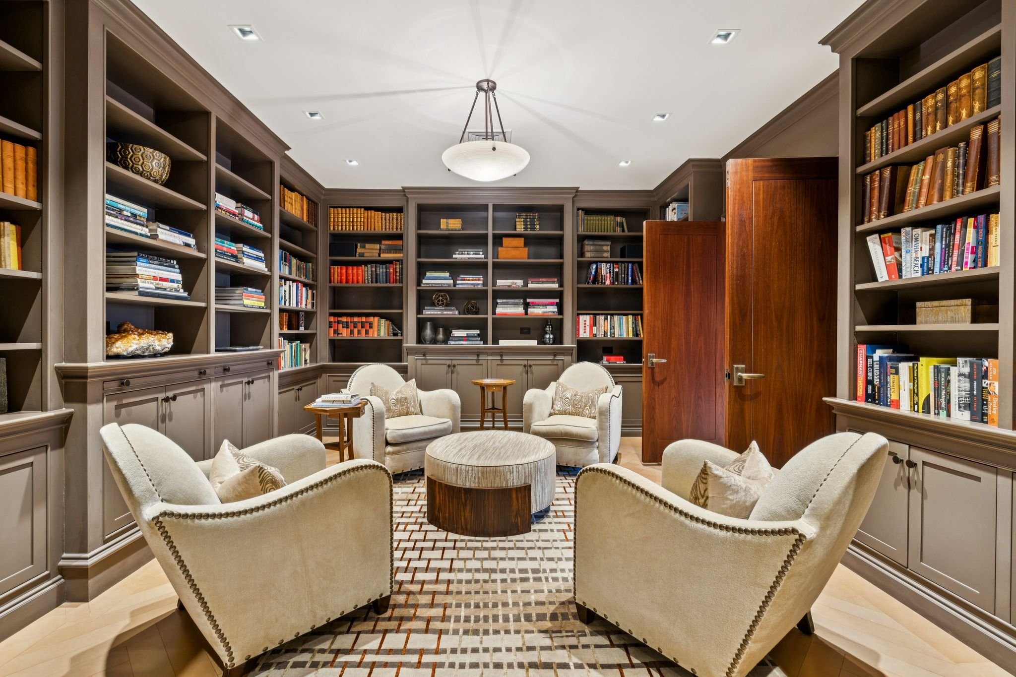 A cozy library or reading room with built-in bookshelves filled with books and decorative items. There are six cream-colored armchairs arranged around a round ottoman. The room features a wooden ceiling light fixture, crown molding, and a patterned rug on light wood flooring.