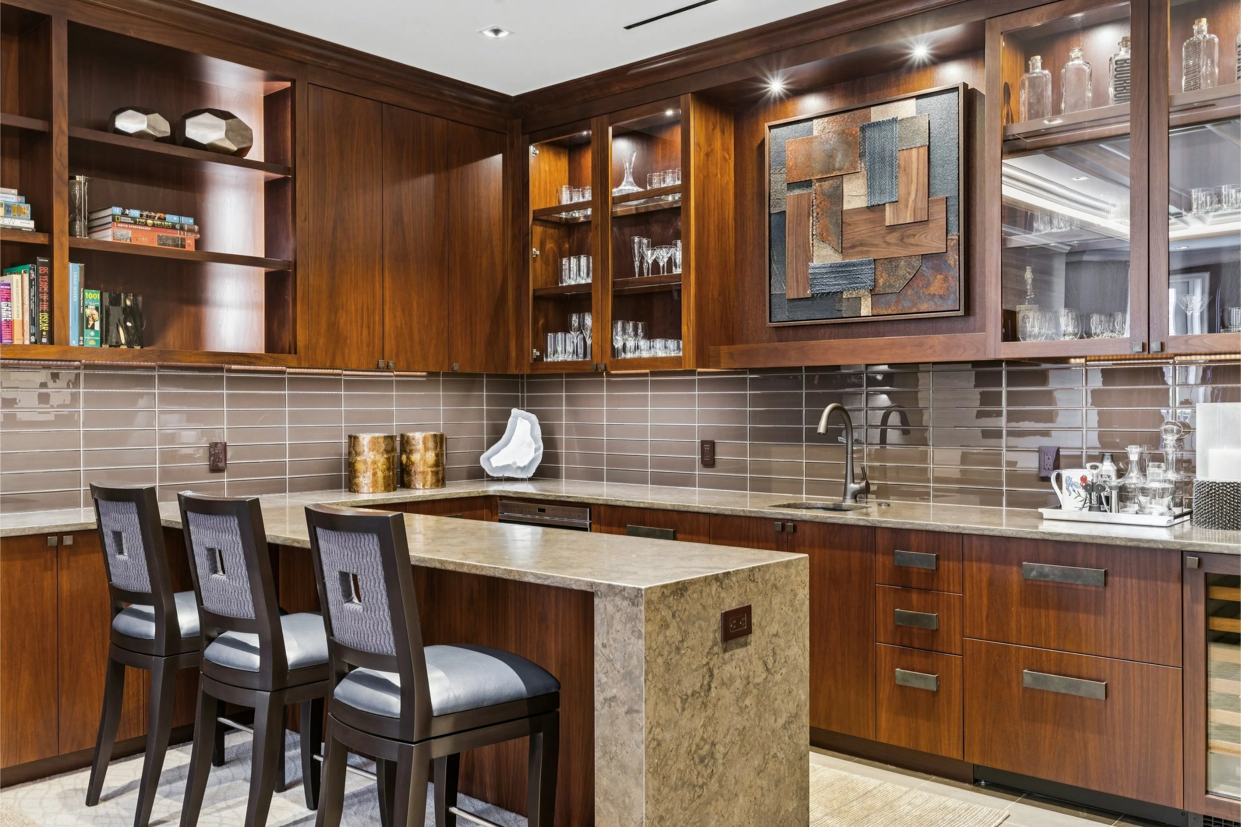 Kitchen with wooden cabinets, open glass shelves, and a beige granite island with bar stools. Glassware and decorative items on shelves, abstract art on wall, and subway tile backsplash.