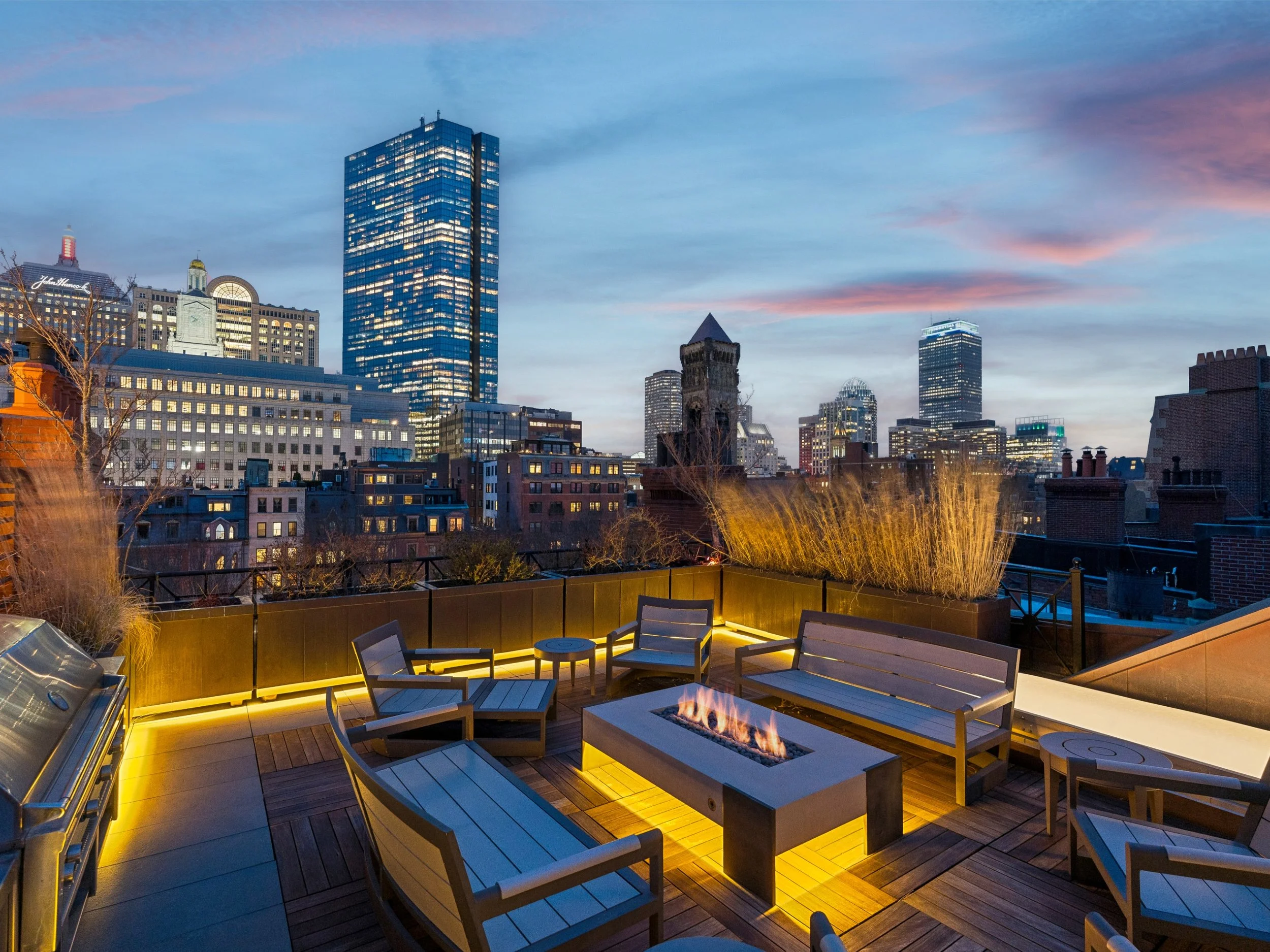 City skyline at dusk with modern high-rise buildings and a rooftop patio with chairs, a fireplace, and a grill.