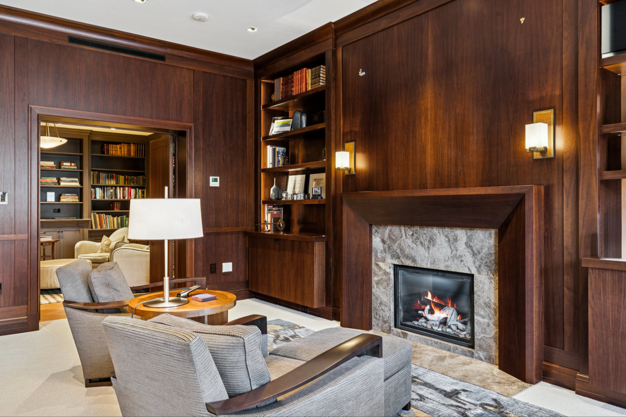 Living room with a wood-paneled wall, built-in bookshelf, and a fireplace with stone surround, lit with wall sconces. There are chairs and a table in the foreground, and a doorway leading to a library or office area with bookshelves in the background