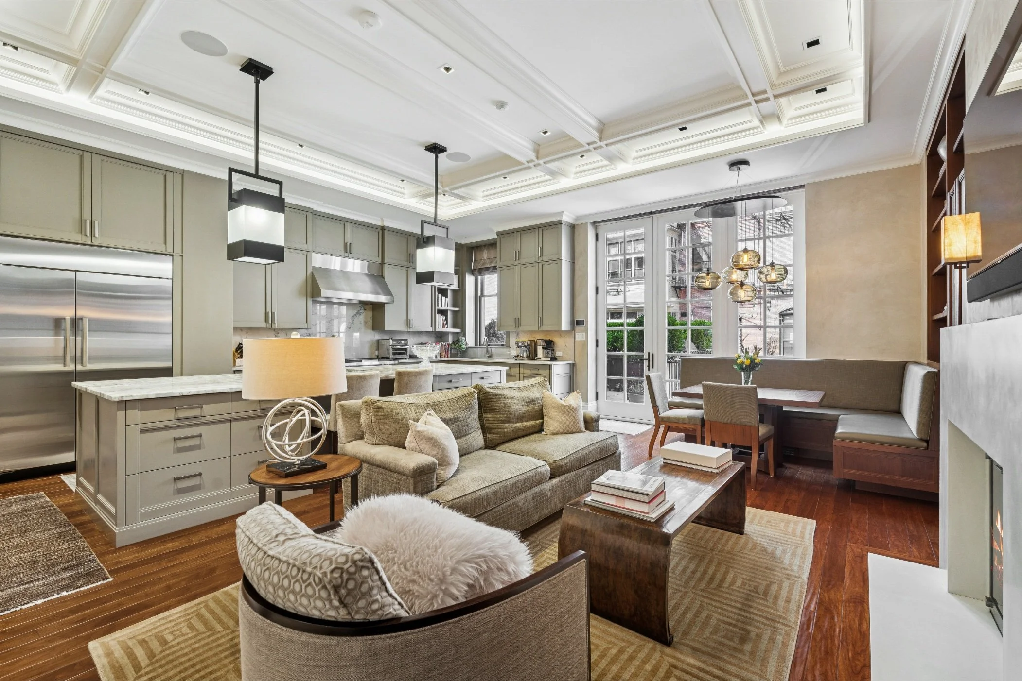 Living room with a beige sofa, armchair with plush white pillow, coffee table, hanging pendant lights, large windows, kitchen area with cabinetry, and wooden flooring.