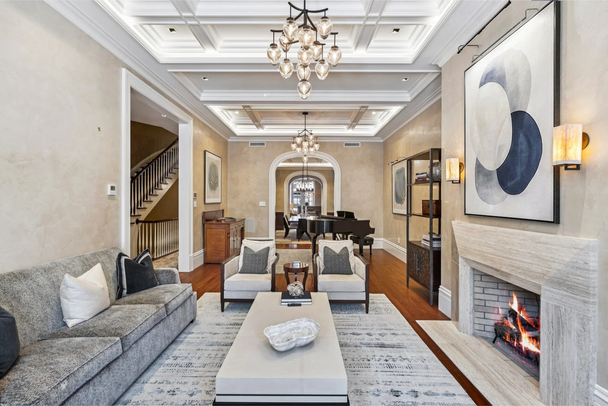 Spacious living room with beige walls, hardwood floors, a gray sofa with pillows, two white armchairs, a coffee table with a decorative bowl, a modern fireplace with flames, abstract wall art, a black grand piano, and a dining area with a chandelier 