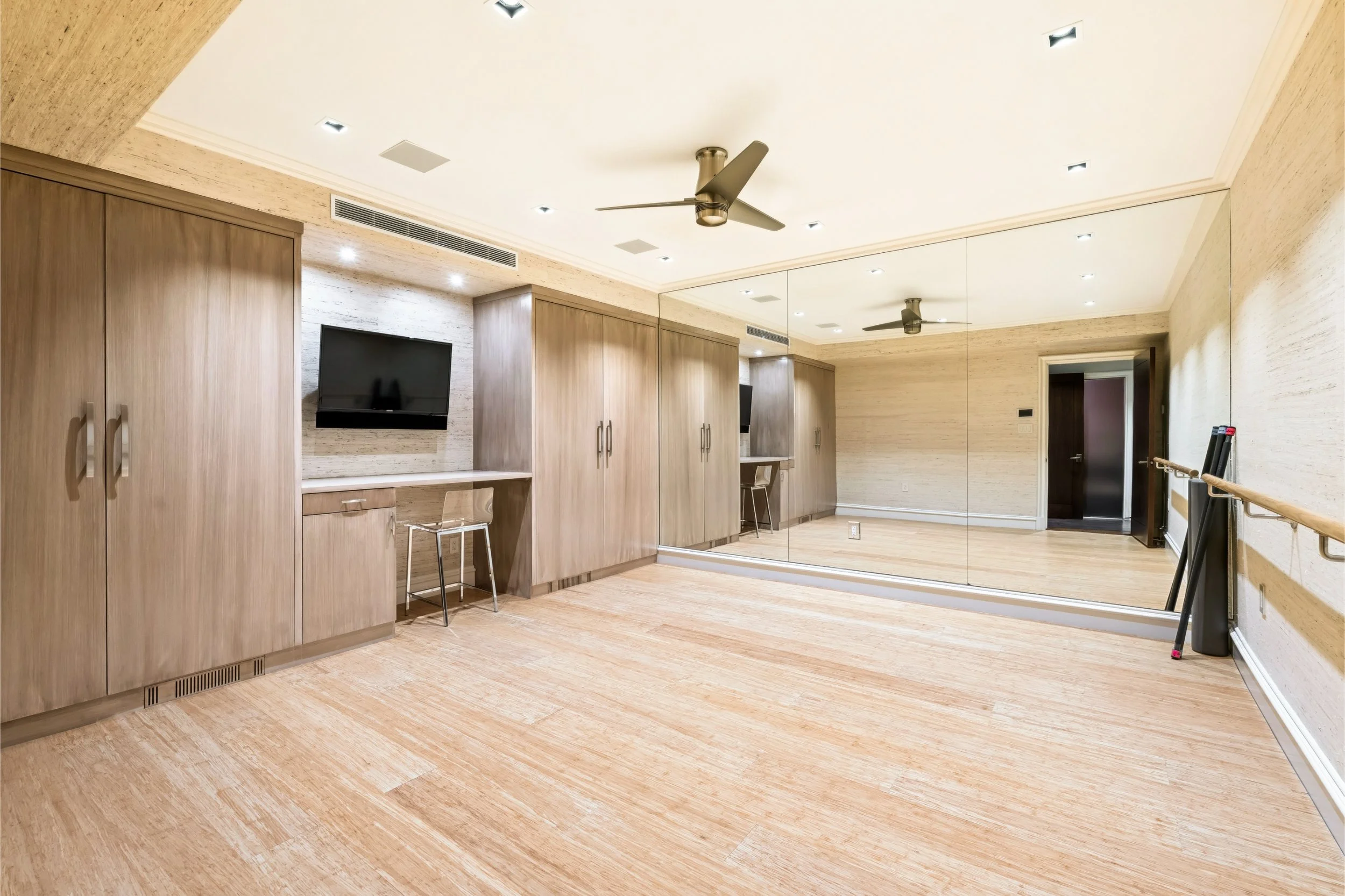 A modern dance or fitness studio with wooden floors, beige walls, mirror panels on one wall, ceiling fans, and recessed lighting.