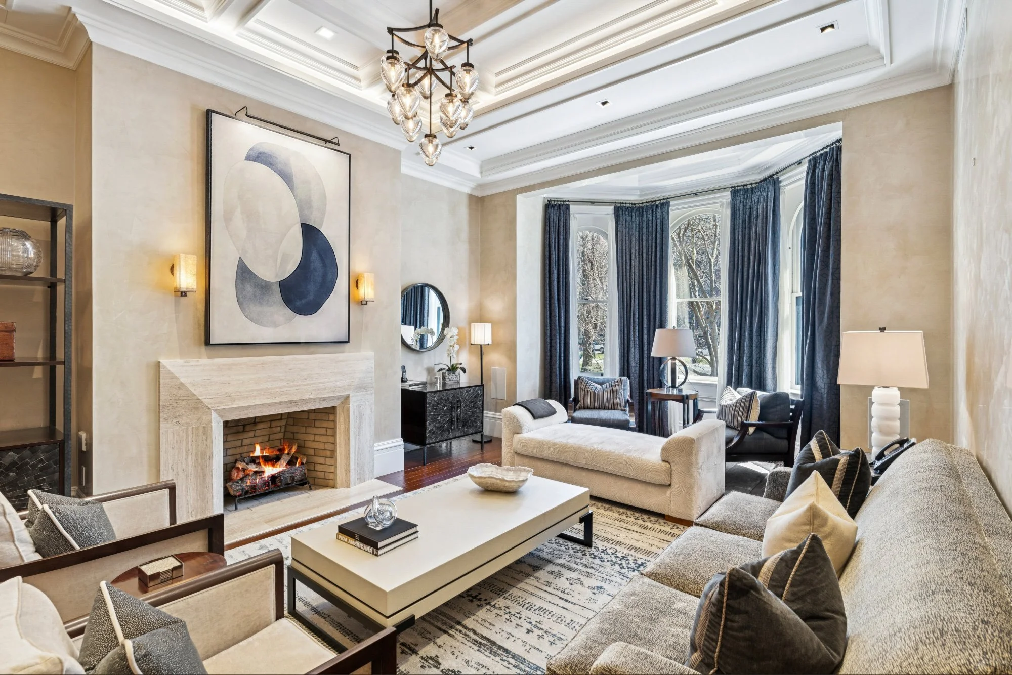 Spacious living room with high ceilings, large windows with blue curtains, a central beige sofa, multiple armchairs, a white coffee table, abstract art above a fireplace, and various lamps and decorative items.