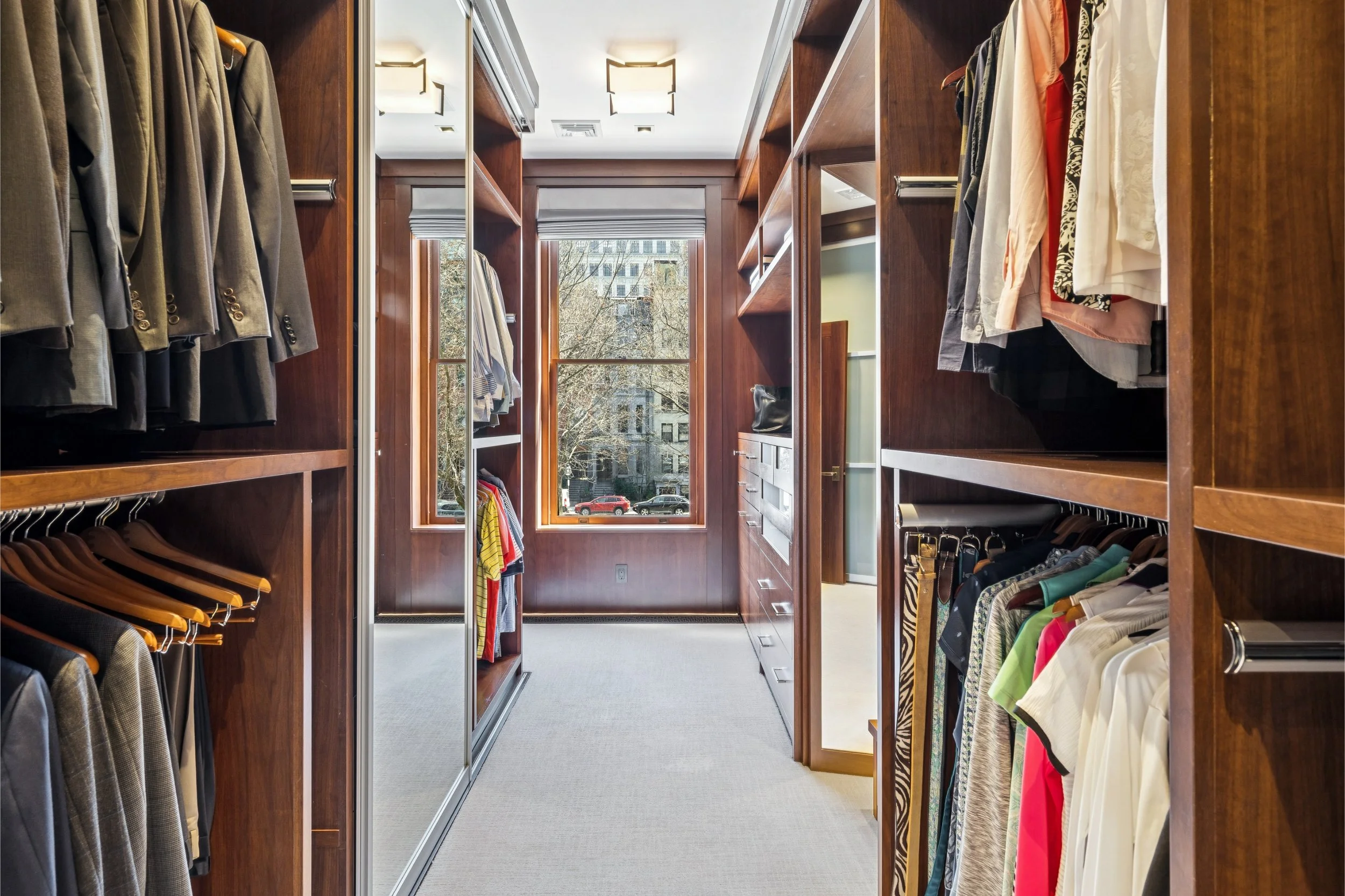A spacious walk-in closet with wooden shelves and hanging rods filled with men's suits, shirts, and pants. A large window at the end of the closet lets in natural light, showing trees and buildings outside.