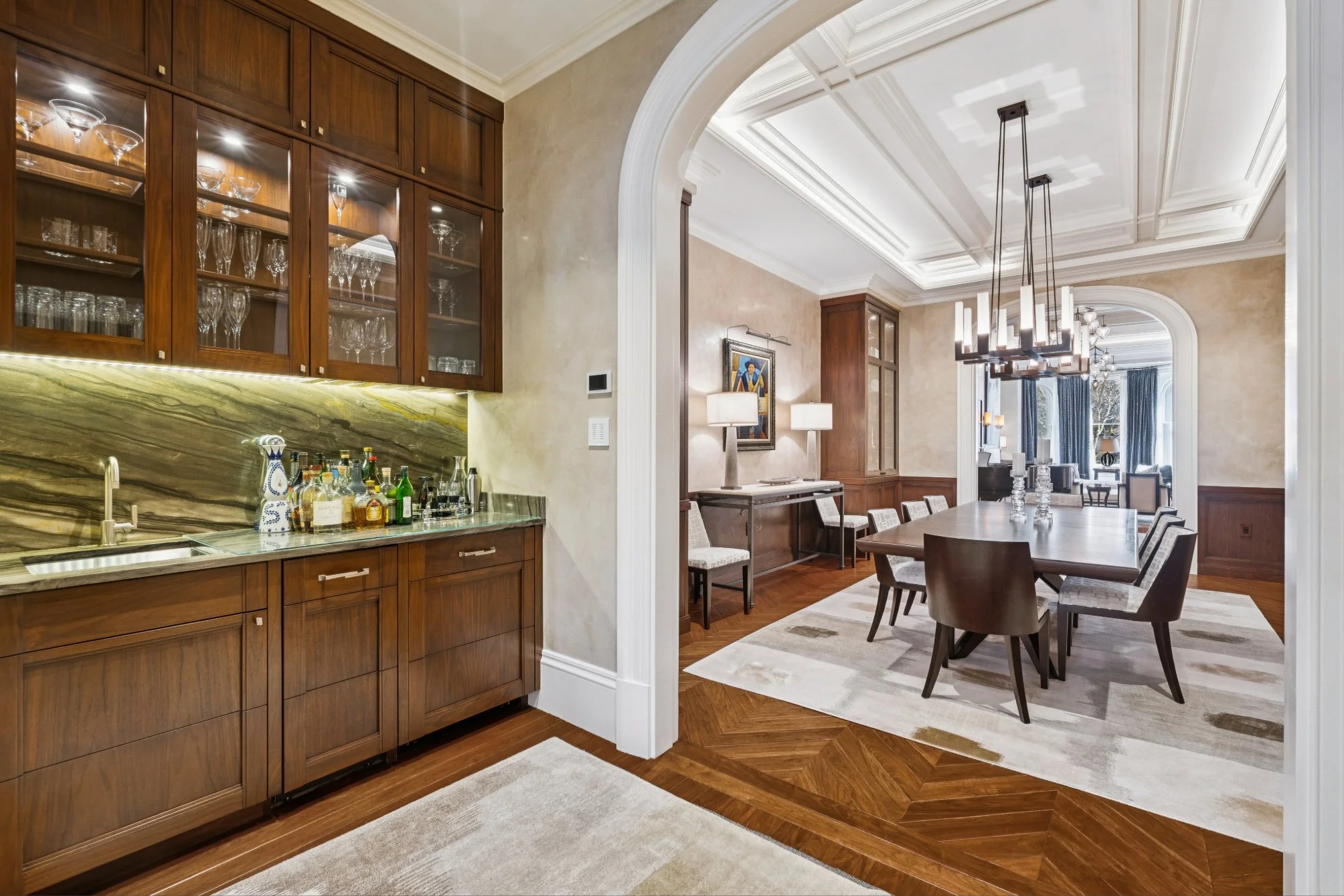 Elegant open-concept living and dining area with warm wood furnishings, a decorated ceiling, and a built-in bar area with glassware, illuminated by modern pendant lights.