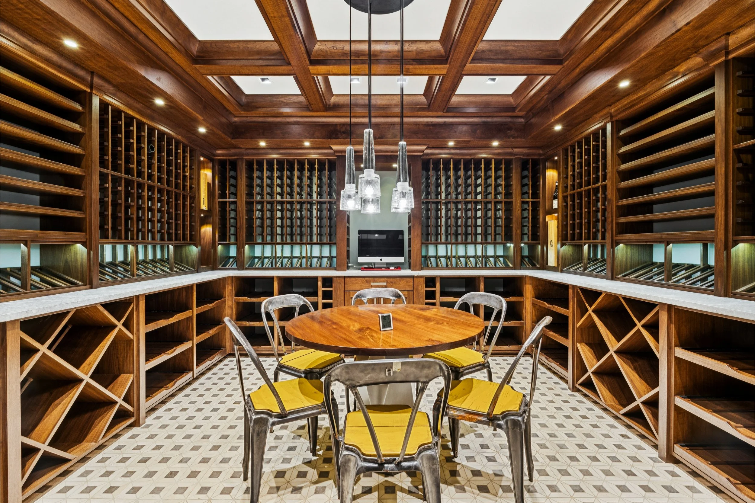 Wood-paneled wine cellar with built-in wine racks, a round table with yellow chairs, pendant lights, and a checkered tile floor.