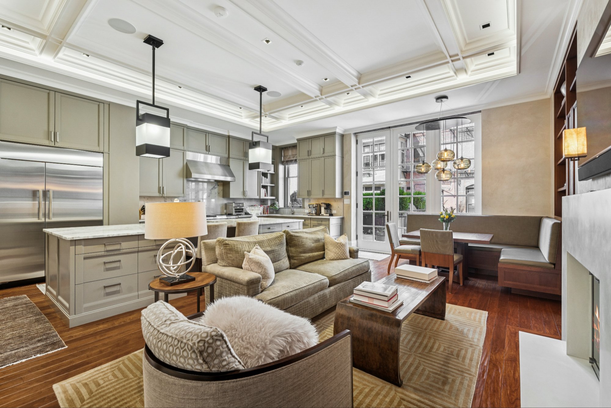 Bright and spacious living room with high ceilings, large windowed doors, a kitchen area, and cozy seating arrangements including a sofa and armchair.