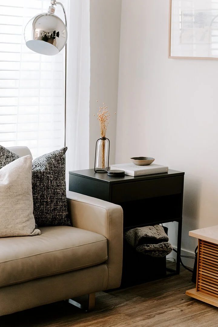 Living room corner with beige sofa, black side table holding a decorative vase with dried flowers and a bowl, a wall-mounted lamp, and white curtains.