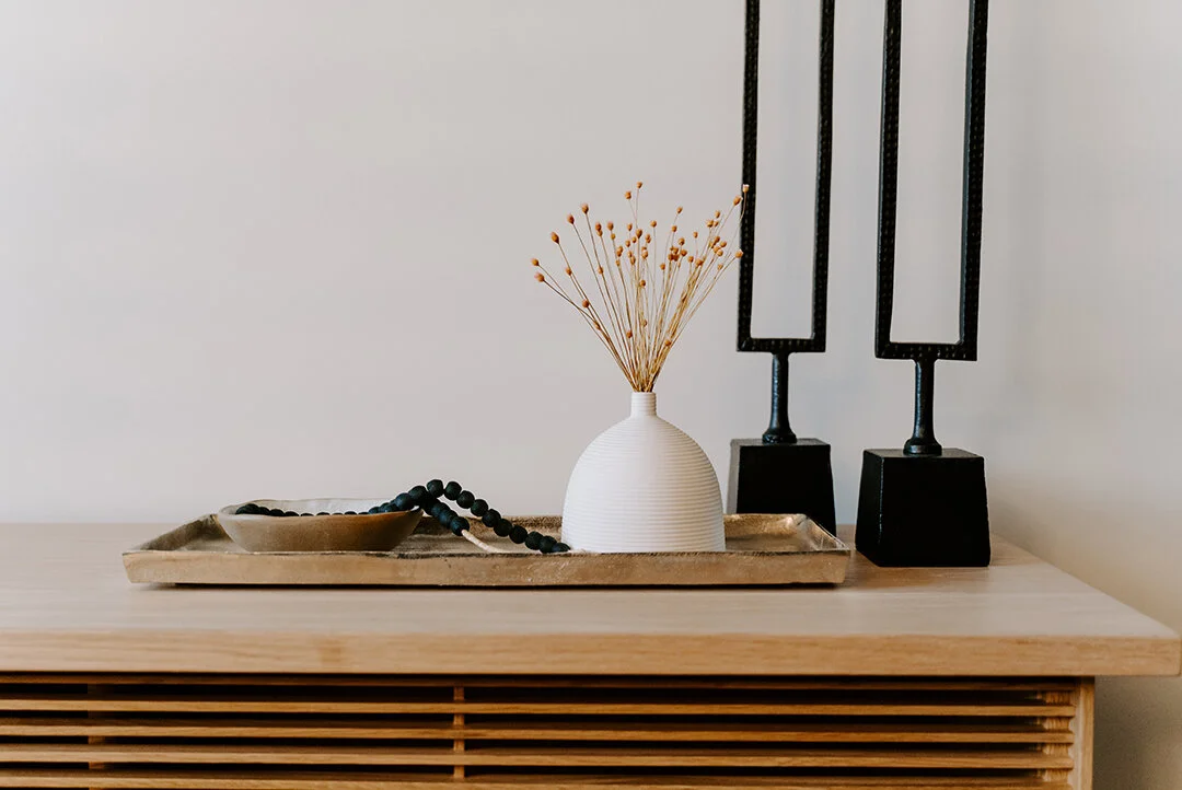 Decorative items on a wooden surface including a white vase with dried flowers, a tray with black beads, and two black geometric sculptures.
