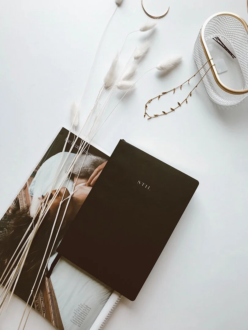 Flat lay of a white surface featuring a black notebook with 'STILL' written on it, magazines, dried white grasses, jewelry including crescent moon and leaf-shaped necklaces, and a clear textured container with a gold rim and a pair of sunglasses inside.