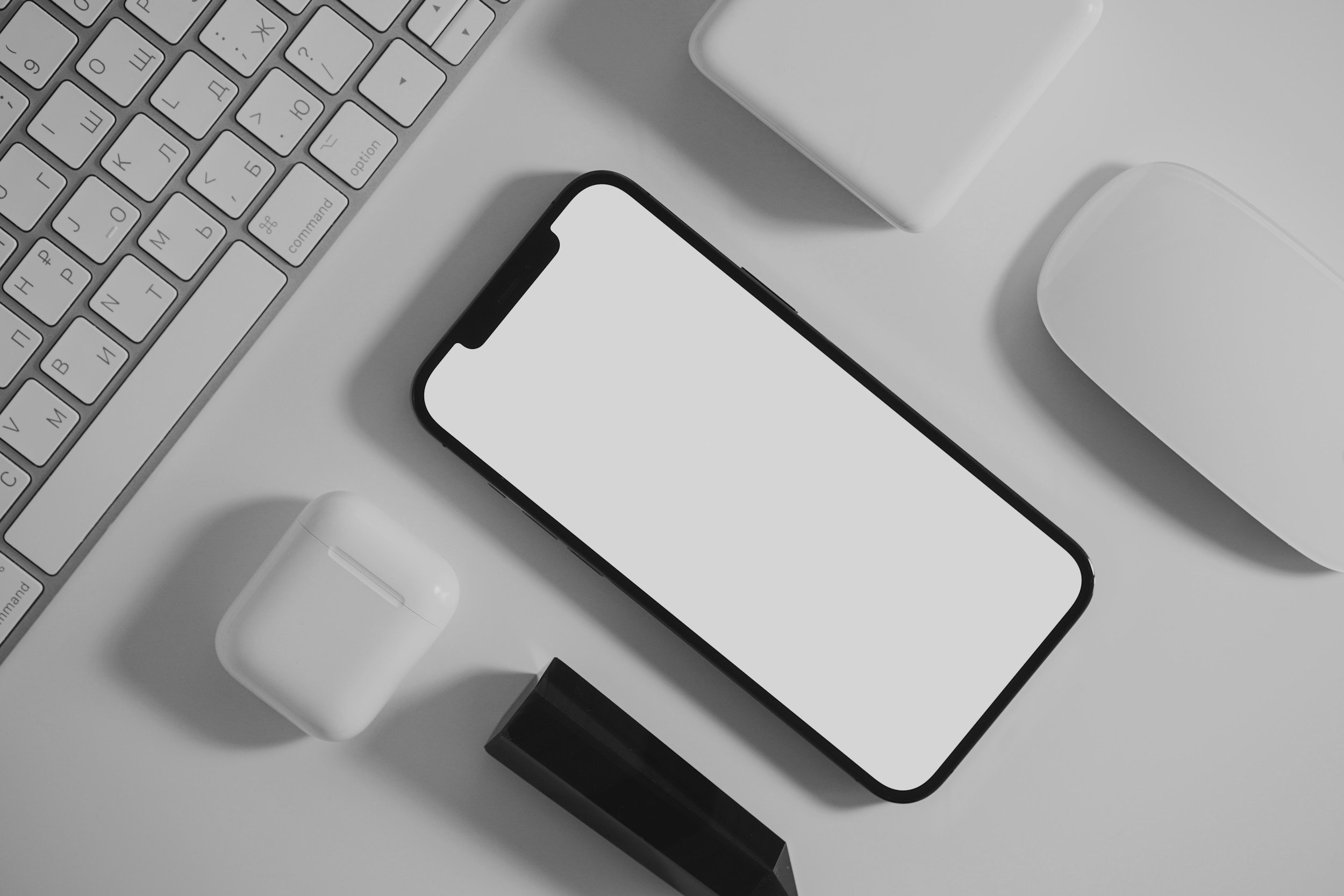 Close-up of a modern smartphone with a blank white screen, alongside a wireless earbud case, a computer keyboard, a computer mouse, and a black rectangular object on a white surface.