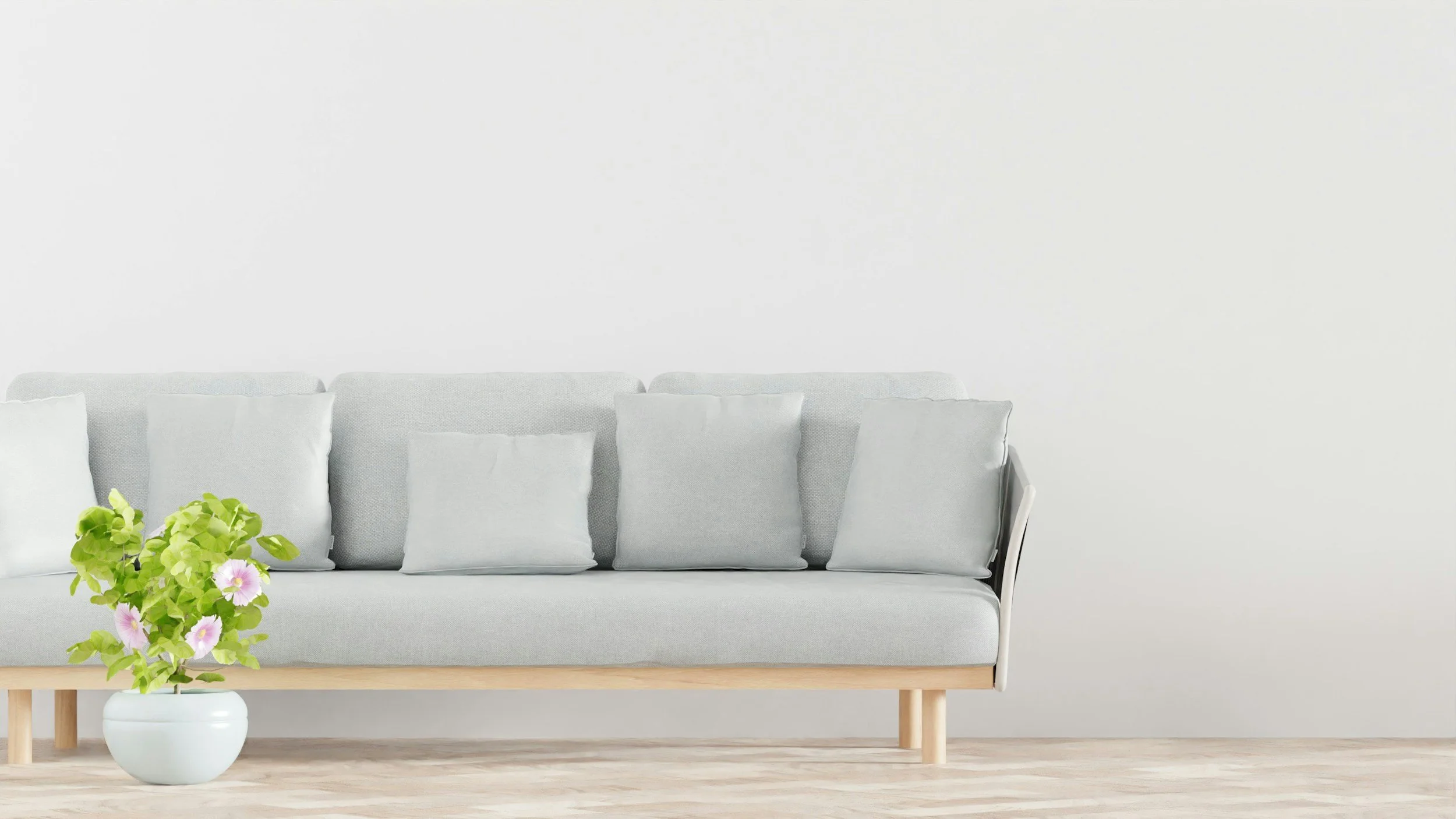 White sofa with multiple white pillows, a light-colored wooden table with a white vase holding green leafy plants and pink flowers, and a plain white wall in the background.