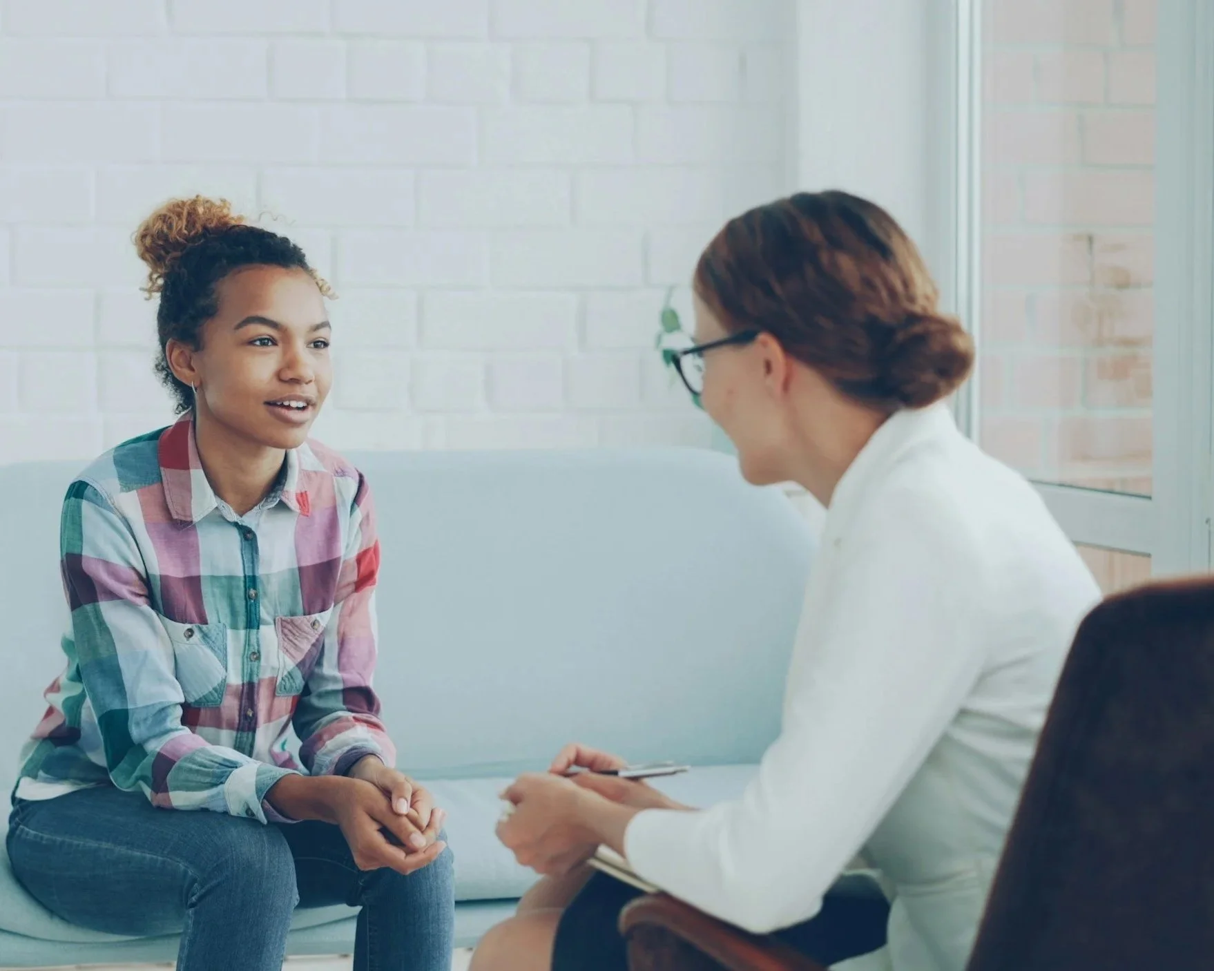 A young woman with curly hair and a plaid shirt sitting and talking to a woman in a white coat and glasses, who is taking notes on a clipboard in a bright, modern room with white brick walls and large windows.