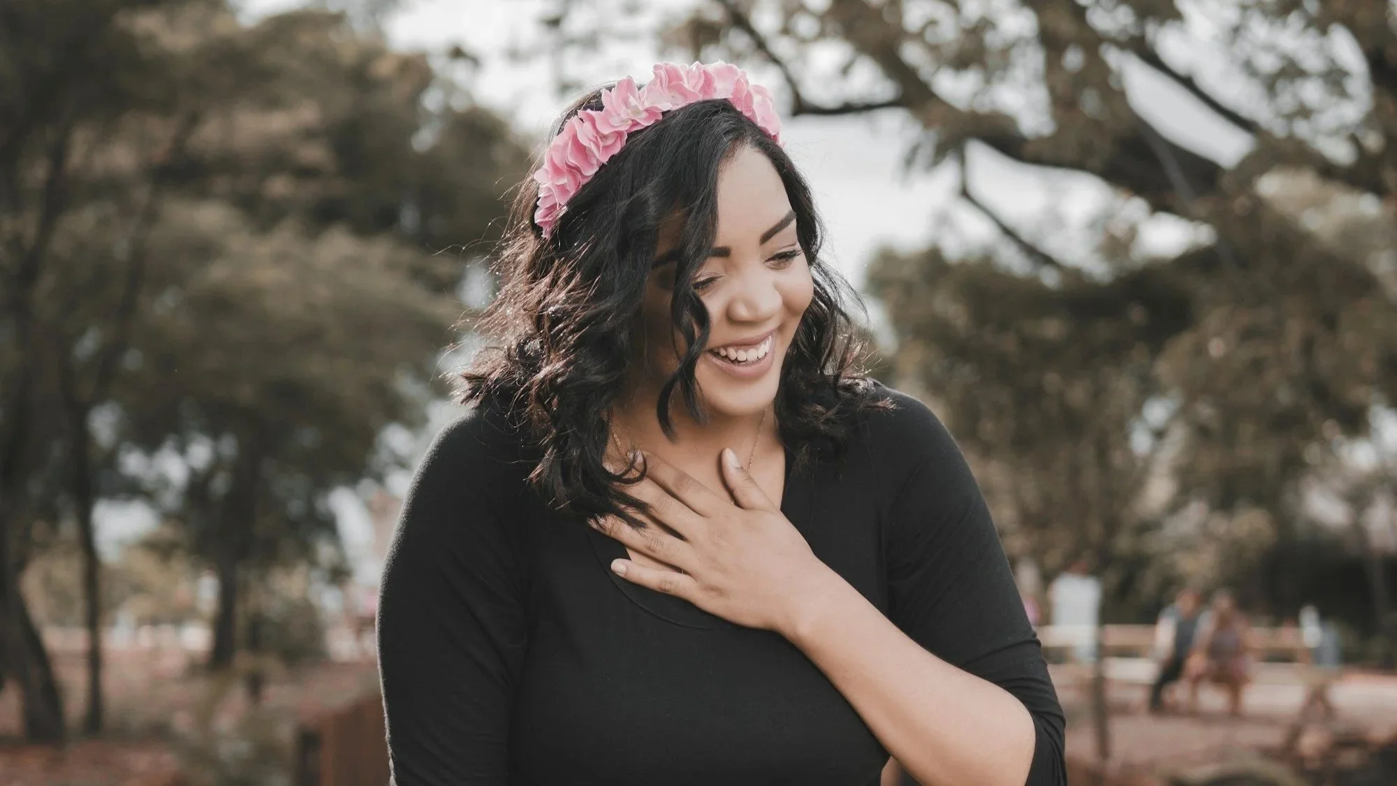 A woman with black, curly hair wearing a pink floral headband and a black top, smiling with her eyes closed, hand on her chest, outdoors in a park with trees in the background.