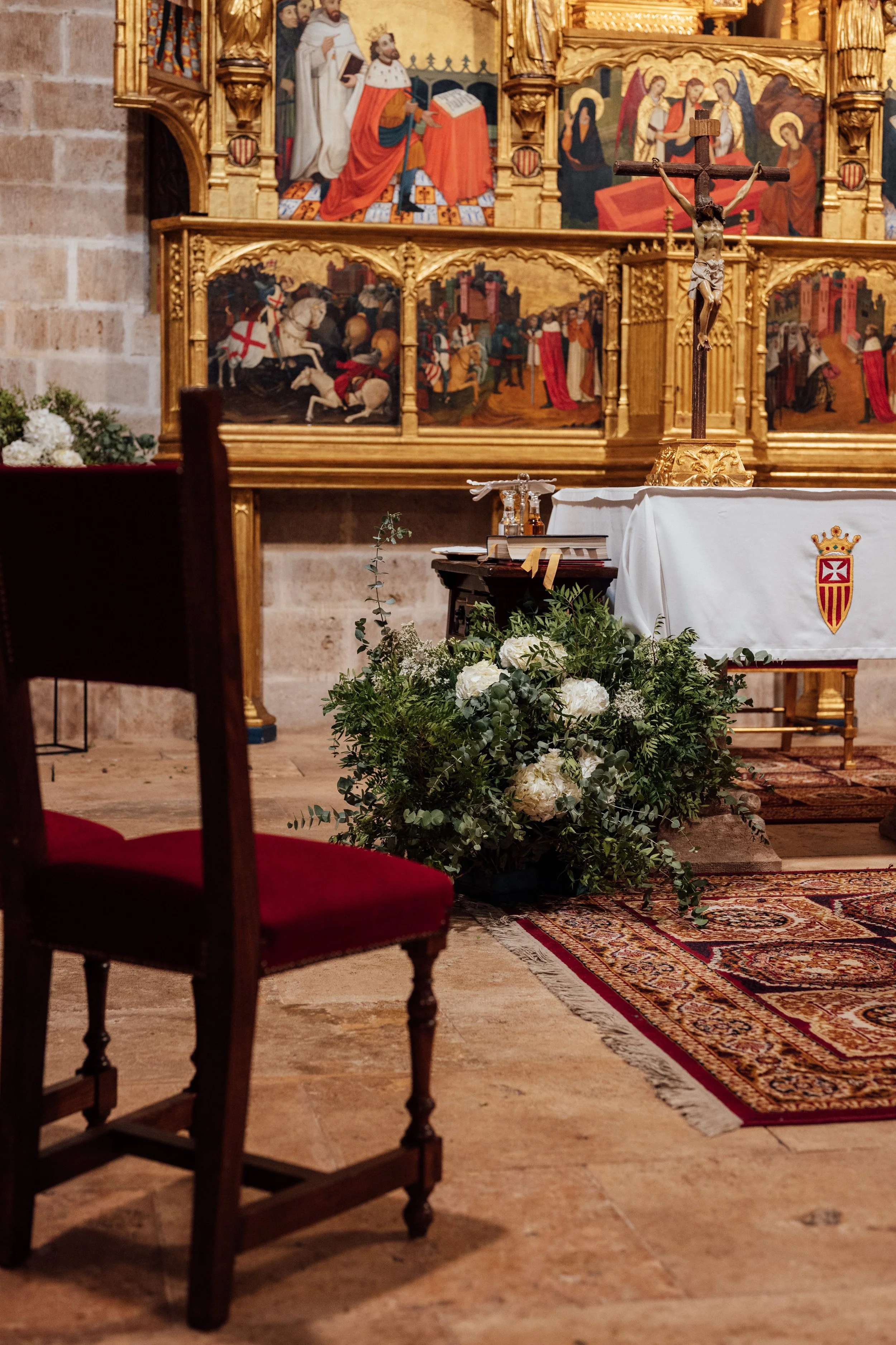 Interior de una iglesia con un altar adornado con un crucifijo, flores y un mantel con escudo, en el fondo hay un retablo dorado con pinturas religiosas.