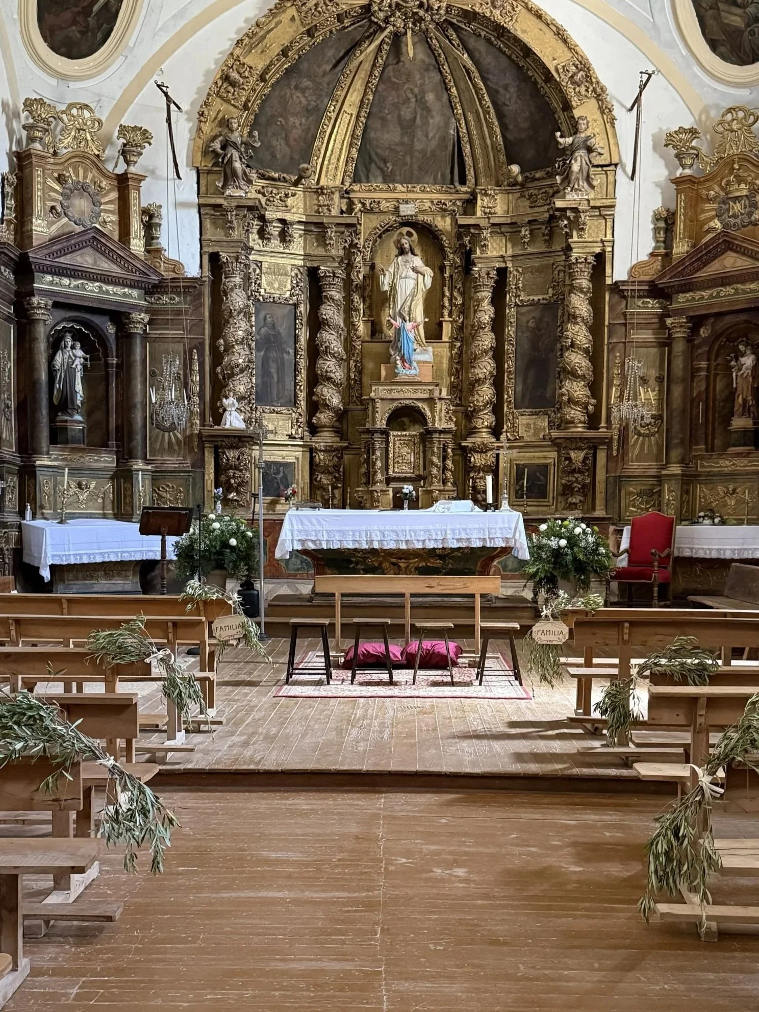Interiores de una iglesia con altar dorado y con imágenes religiosas, decoraciones para una ceremonia, con sillas y bancos de madera, y plantas decorativas.