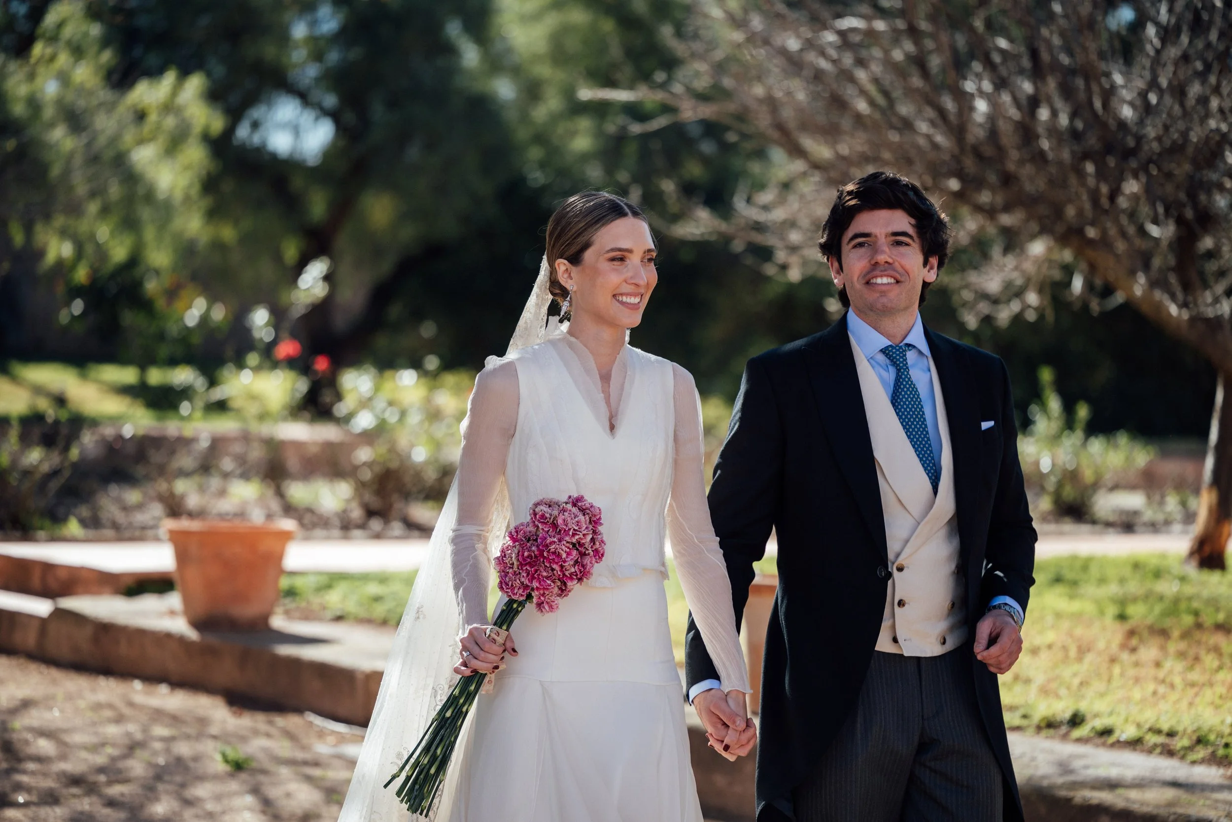 Pareja de recién casados caminando en un parque, la novia lleva un vestido blanco y sostiene un ramo de flores rosadas, el novio viste un traje oscuro con una bufanda blanca. Ambos parecen felices y sonrientes.