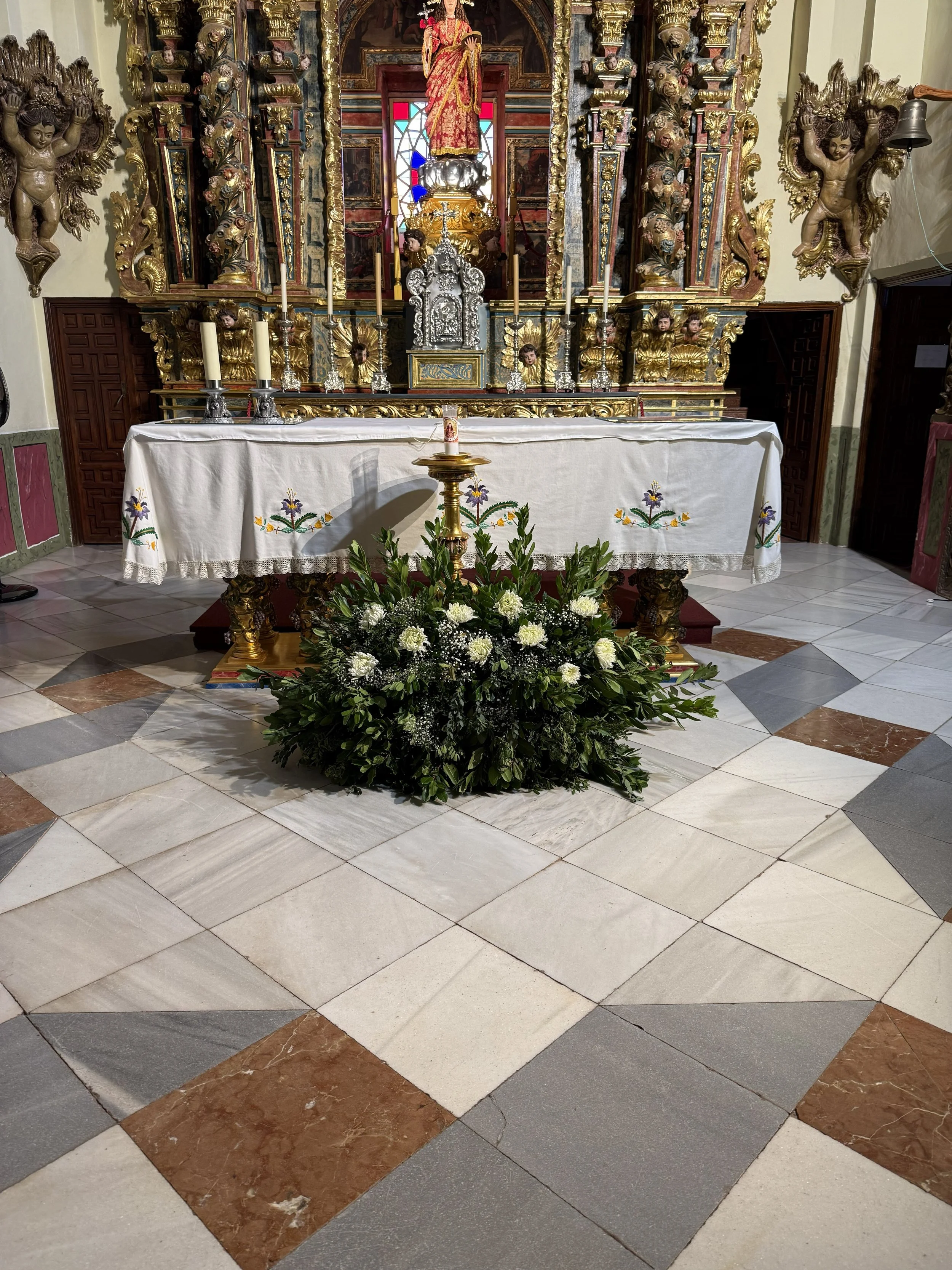 Altar de iglesia con escultura de una figura religiosa en el centro, decorado con velas y flores, con paredes decoradas con relieves y columnas ornamentadas.