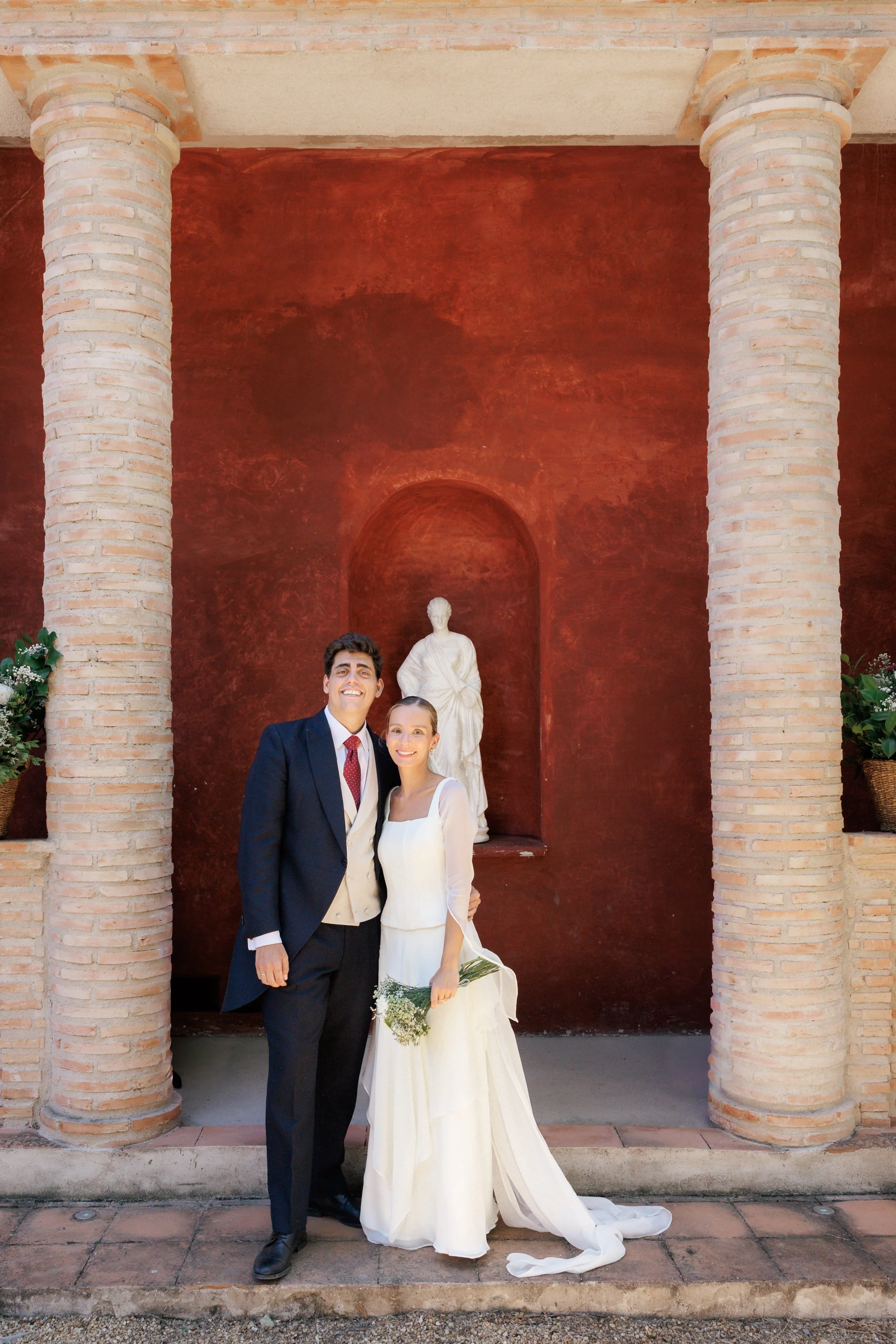 Una pareja de recién casados posa frente a una estructura de ladrillos y una estatua de mármol en un escenario al aire libre. La mujer lleva un vestido de novia largo y sostiene un ramo de flores, mientras que el hombre viste un traje oscuro con corbata. Ambos sonríen a la cámara.