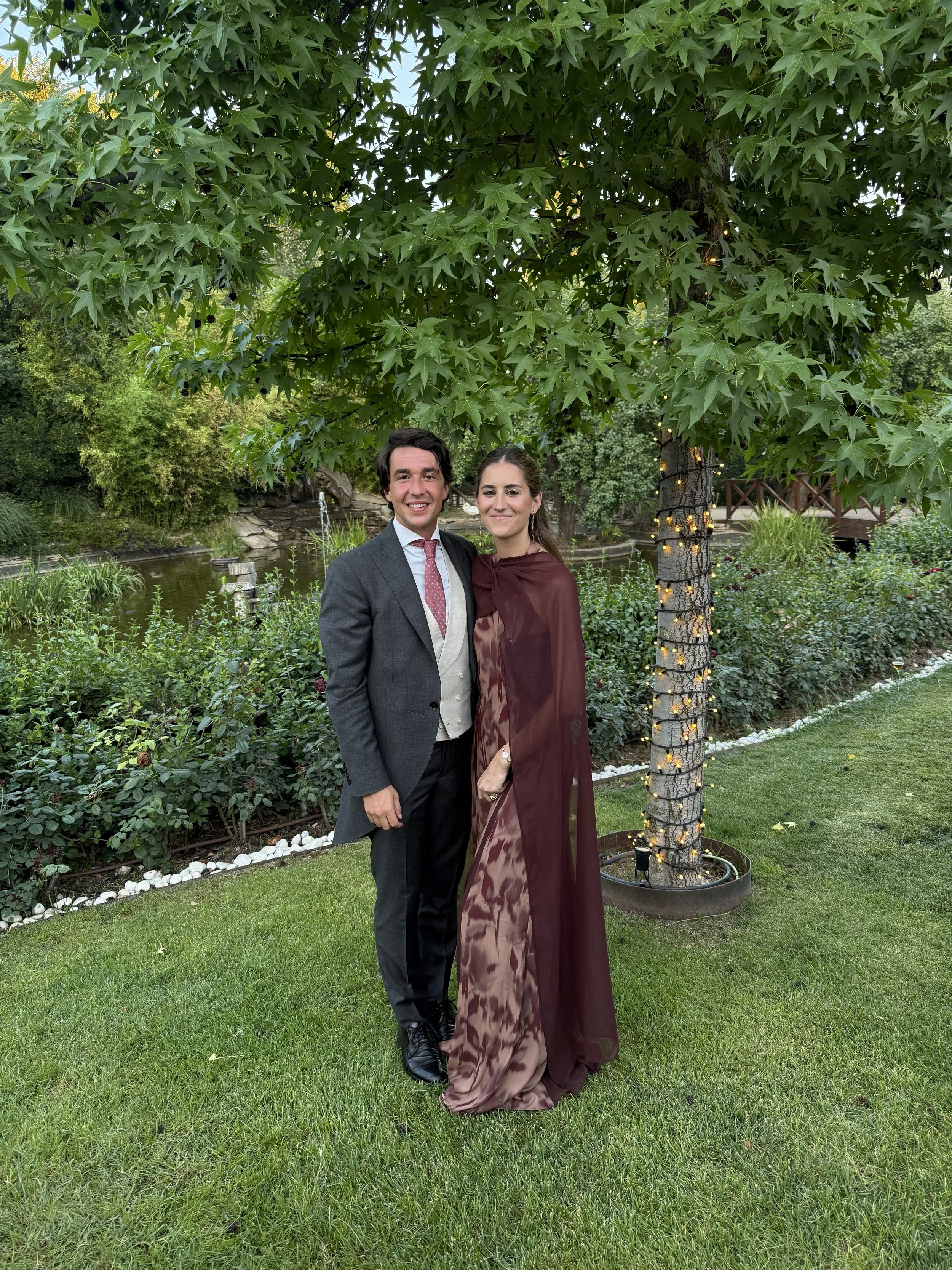 Pareja vestida formalmente, hombre con saco gris y mujer con vestido largo color vino, posando en jardín con árboles y césped, iluminación decorativa en árbol.