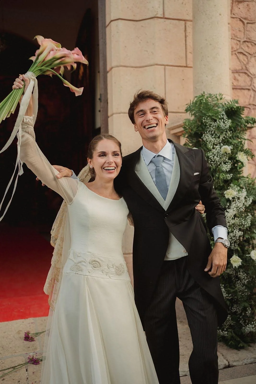 Novios felices en su boda, la novia sostiene un ramo y ambos sonríen, en la entrada de una iglesia decorada con flores.