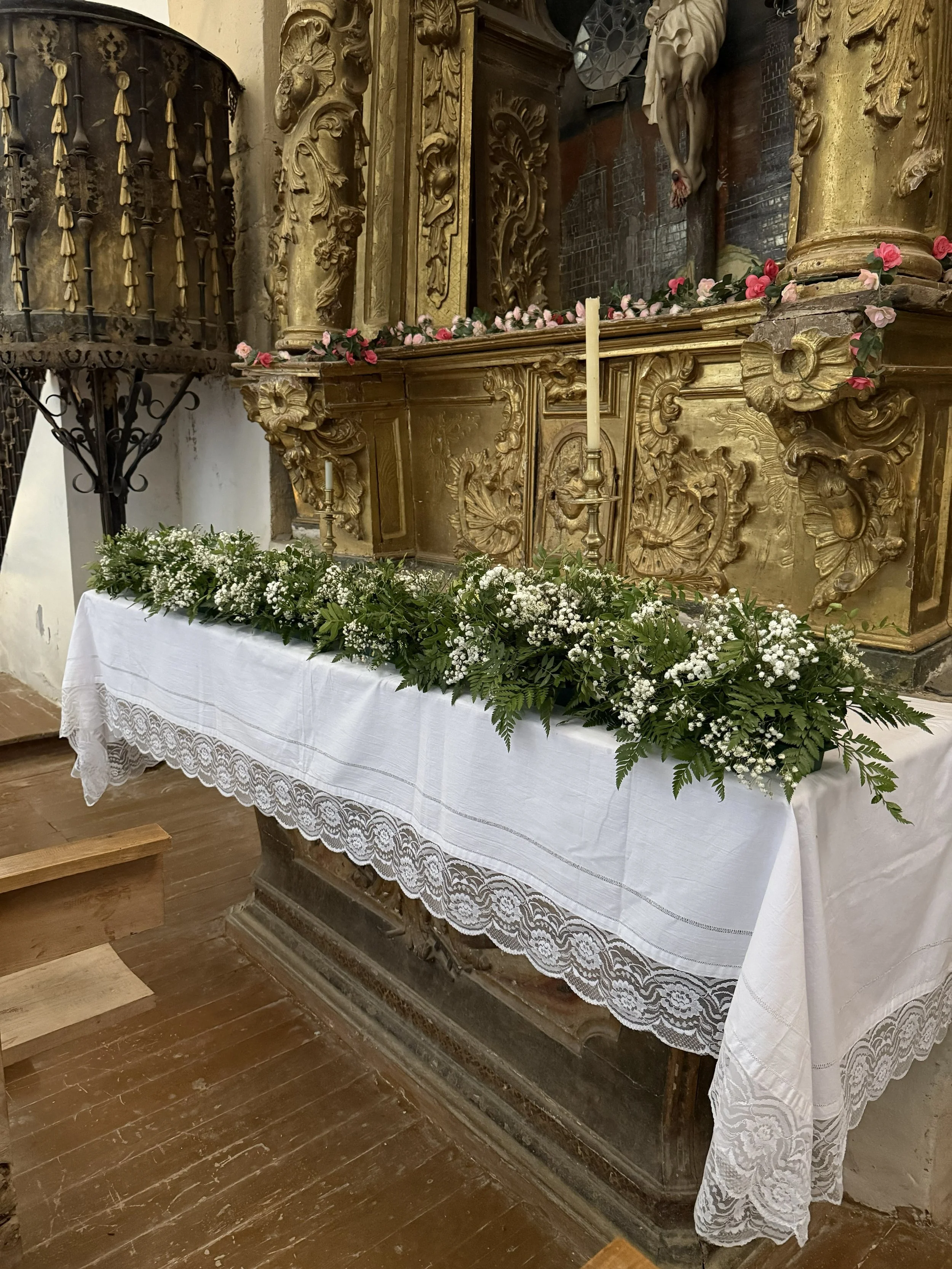 Altar decorativo con flores blancas y verdes, candeleros de cera, en un entorno religioso antiguo con detalles dorados y tallados en madera.