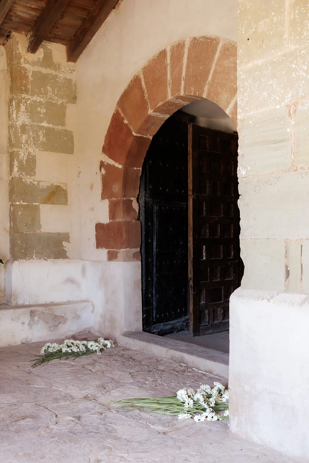 Entrada de una iglesia antigua con arco de piedra y puertas de madera negras, con flores blancas en el suelo.