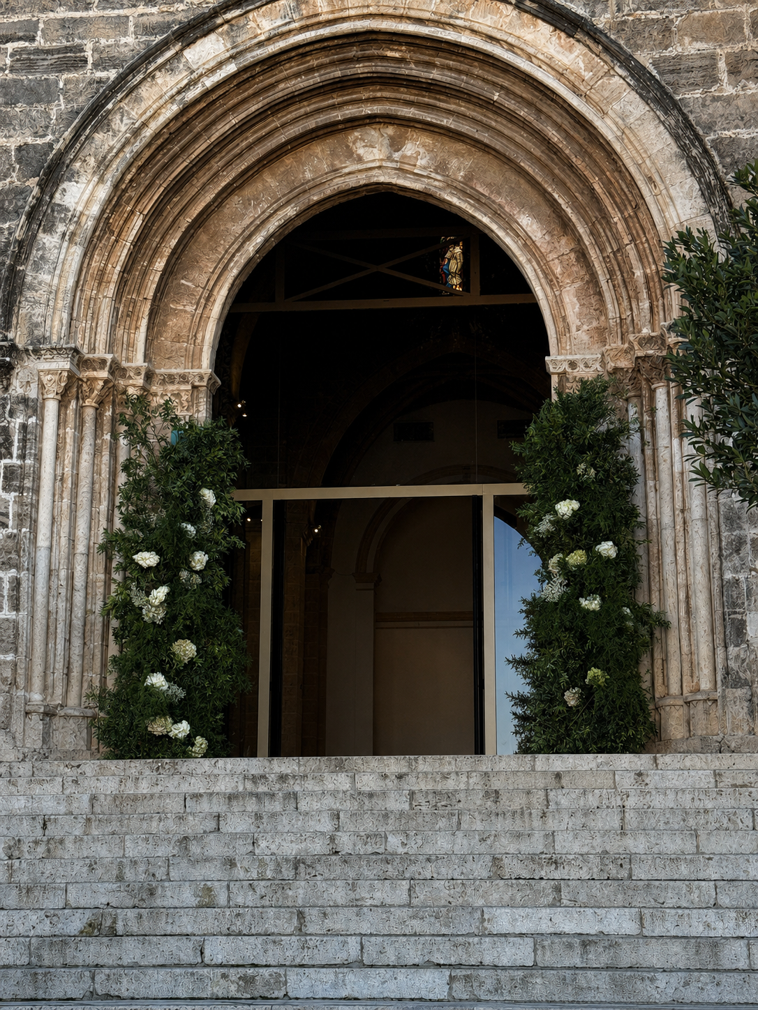 Entrada de una iglesia con arco en piedra y escaleras de piedra, decorada con plantas verdes y flores blancas a ambos lados de la puerta.