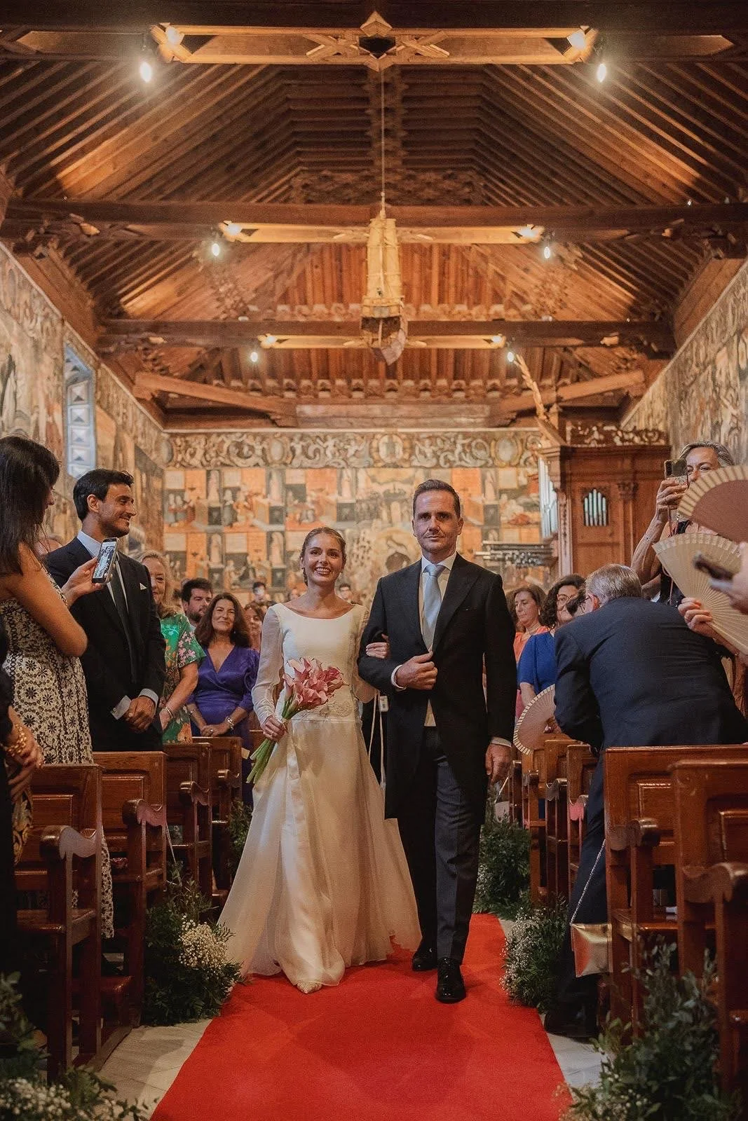 Pareja de boda caminando por pasillo de iglesia, rodeados de invitados, con paredes decoradas con arte y techo de madera, en una ceremonia formal.