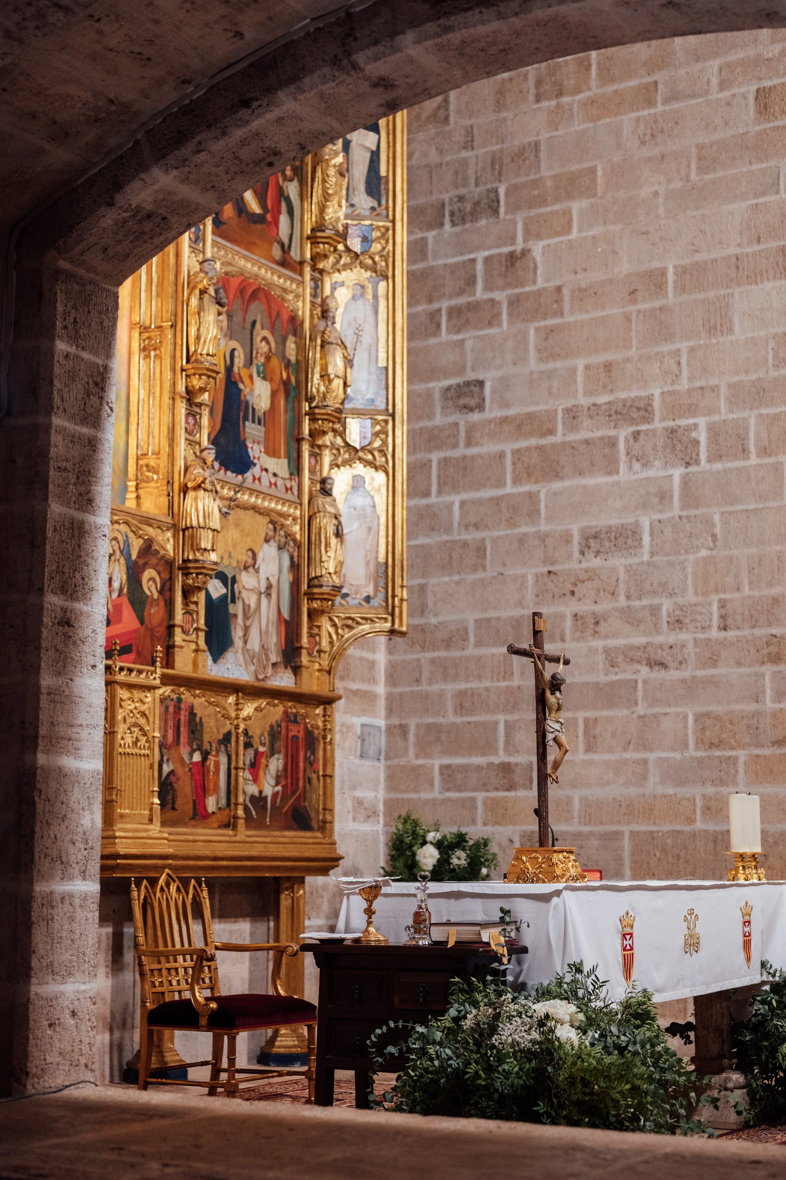 Interior de iglesia con altar adornado con una cruz, flores blancas y manteles blancos con escudos y símbolos religiosos, en un muro de ladrillos y una escultura reliquia de una crucifixión.